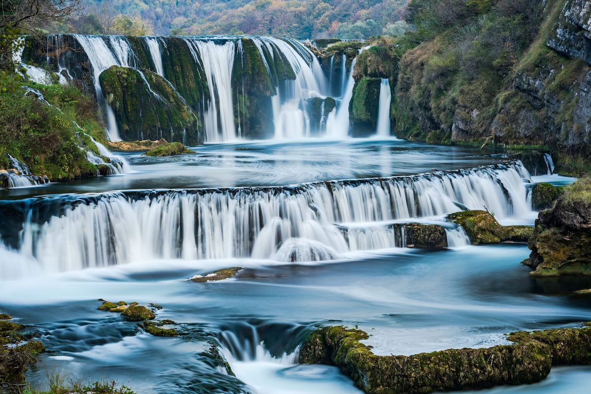 Multi-tiered waterfall cascading over rocky ledges with misty blue water in natural setting.
