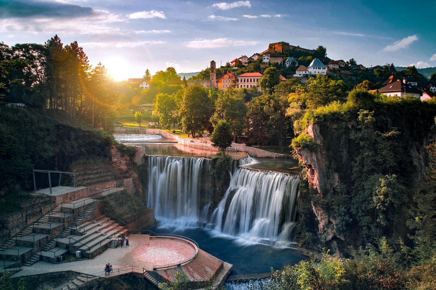 Scenic waterfall cascades through town at sunset with homes perched on cliffs above.