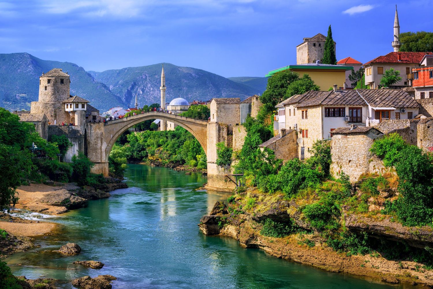 Historic stone bridge spans emerald river in ancient European town with mountain backdrop.