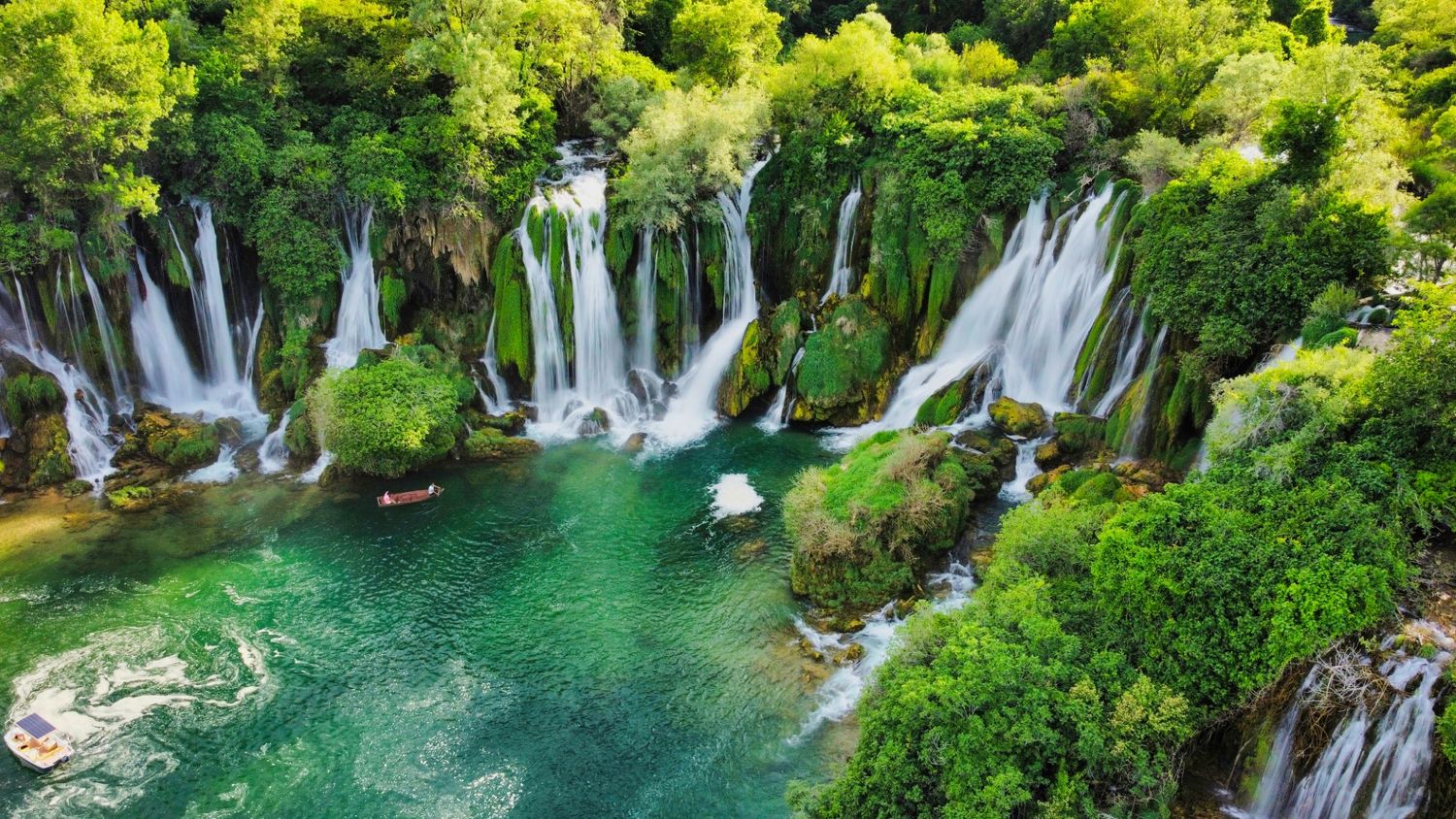 Multiple waterfalls pour into turquoise pool surrounded by lush green forest.