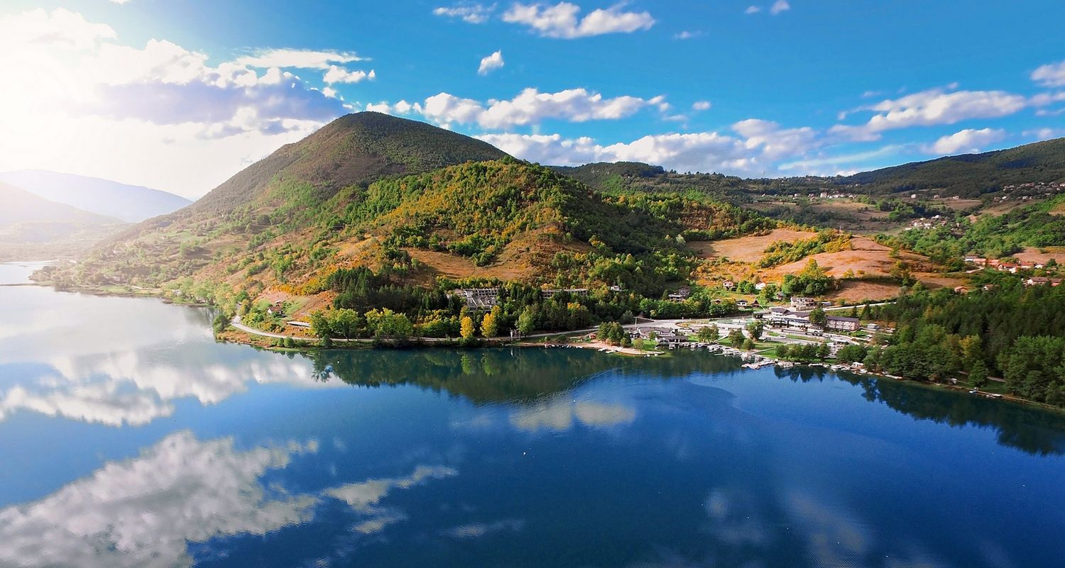 Stunning mountain lake reflecting blue skies and clouds with small village visible along shoreline.