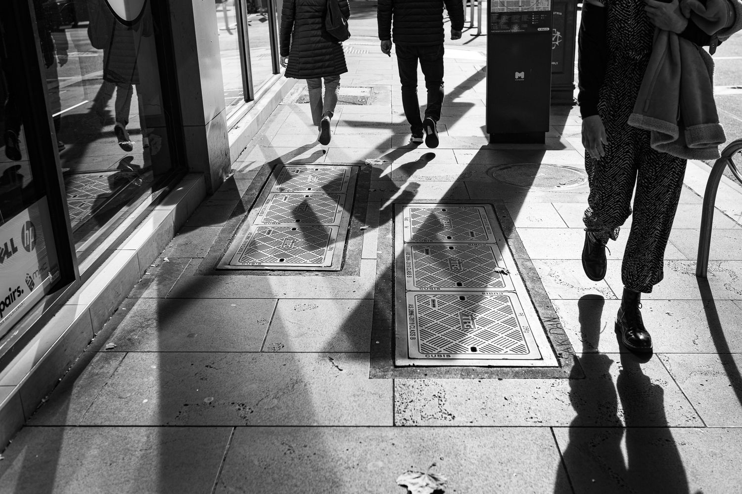 Black and white street photography image showing silhouettes of people walking on a city street.
