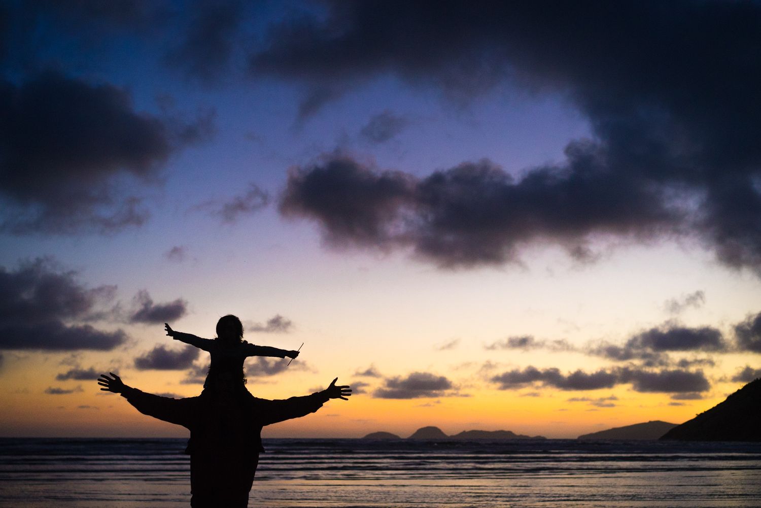 Silhouetted figure stands with outstretched arms against a dramatic sunset sky over ocean horizon.