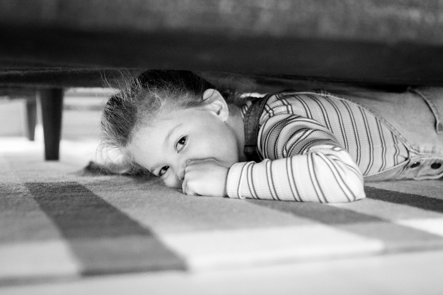 Black and white photograph showing low-angle perspective of a child playing hide and seek under furniture.
