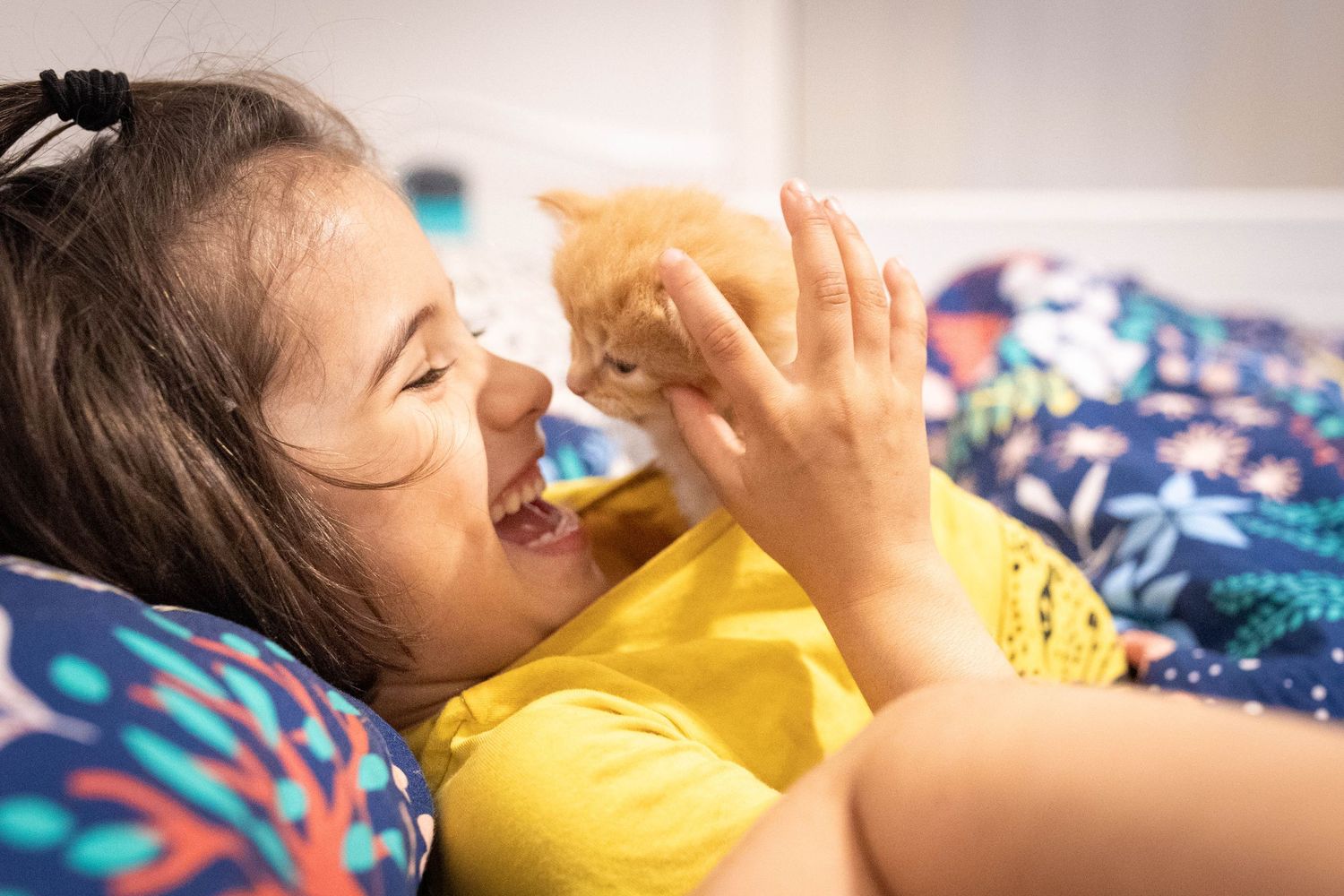 Someone wearing a yellow shirt lies on colorful bedding while playing with an orange cat.