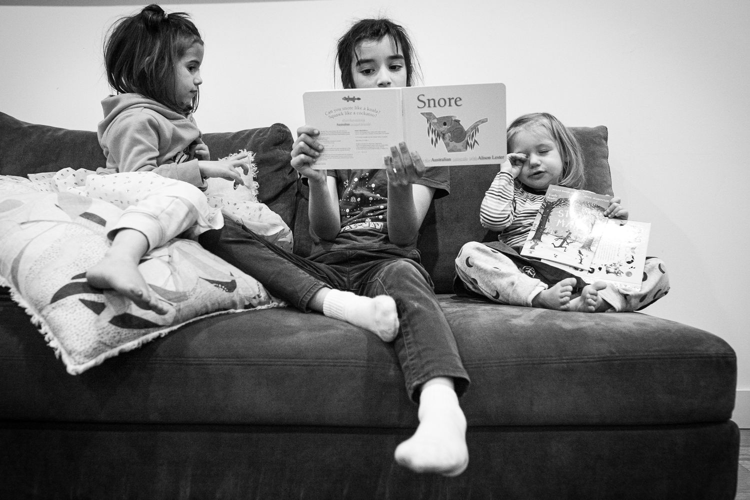 Three children sit together on a couch reading books in this vintage black and white photograph.