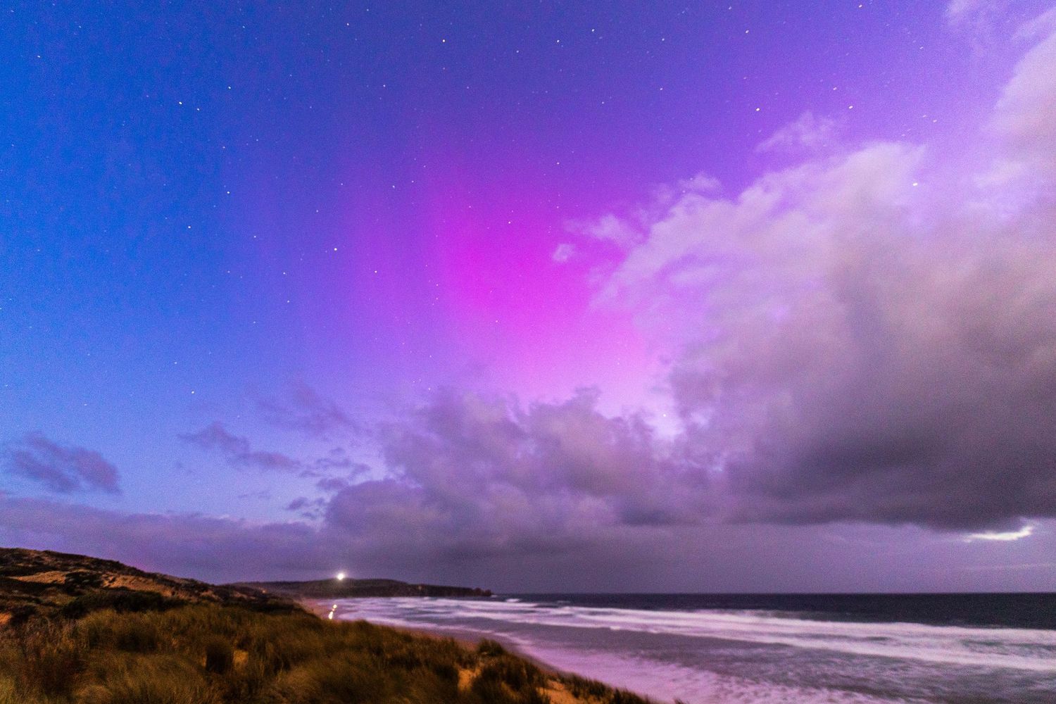 Dramatic pink aurora australis with clouds rolling across a coastal landscape at night.