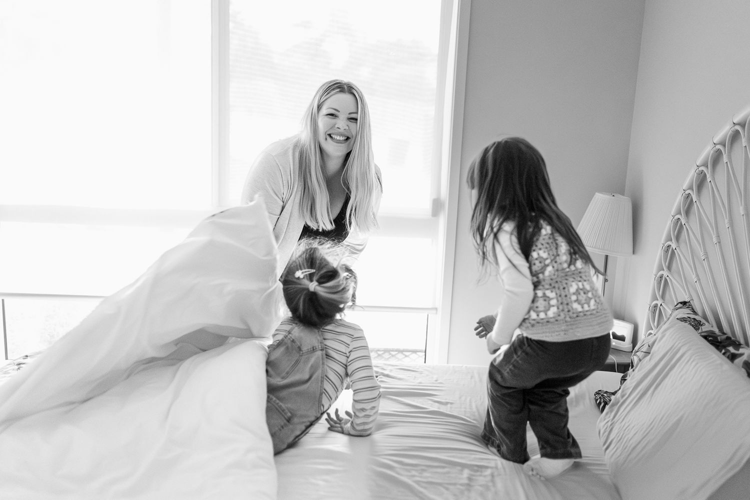 Black and white photo showing playful family moment of surprise on a bed with soft natural lighting through windows.