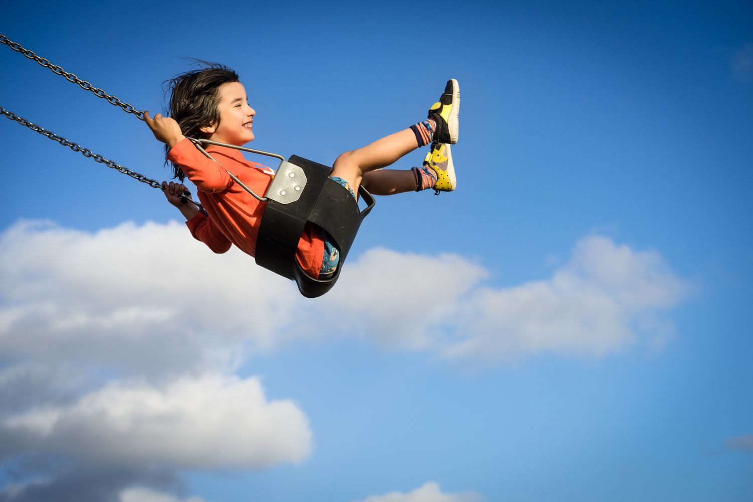 A person swinging on a playground swing against a blue sky with white clouds.