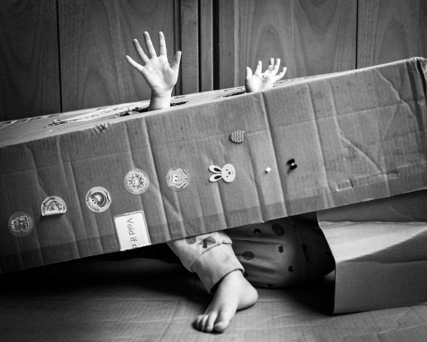 Playful black and white image of a young child emerging from a box with their hands and feet.