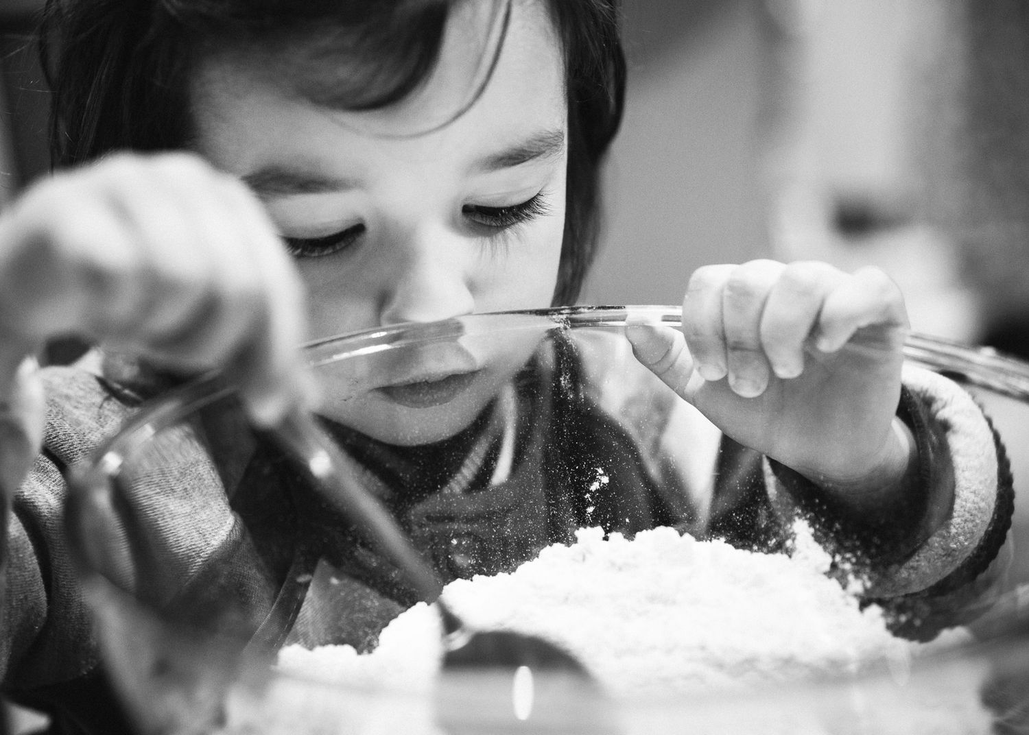 Black and white photo of a small child concentrating while eating a bowl of food with utensils.