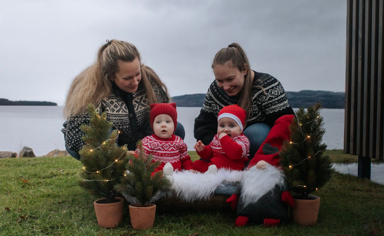 To kvinner og to babyer i juleantrekk fotografert sammen med små juletrær med lys ved vannet.