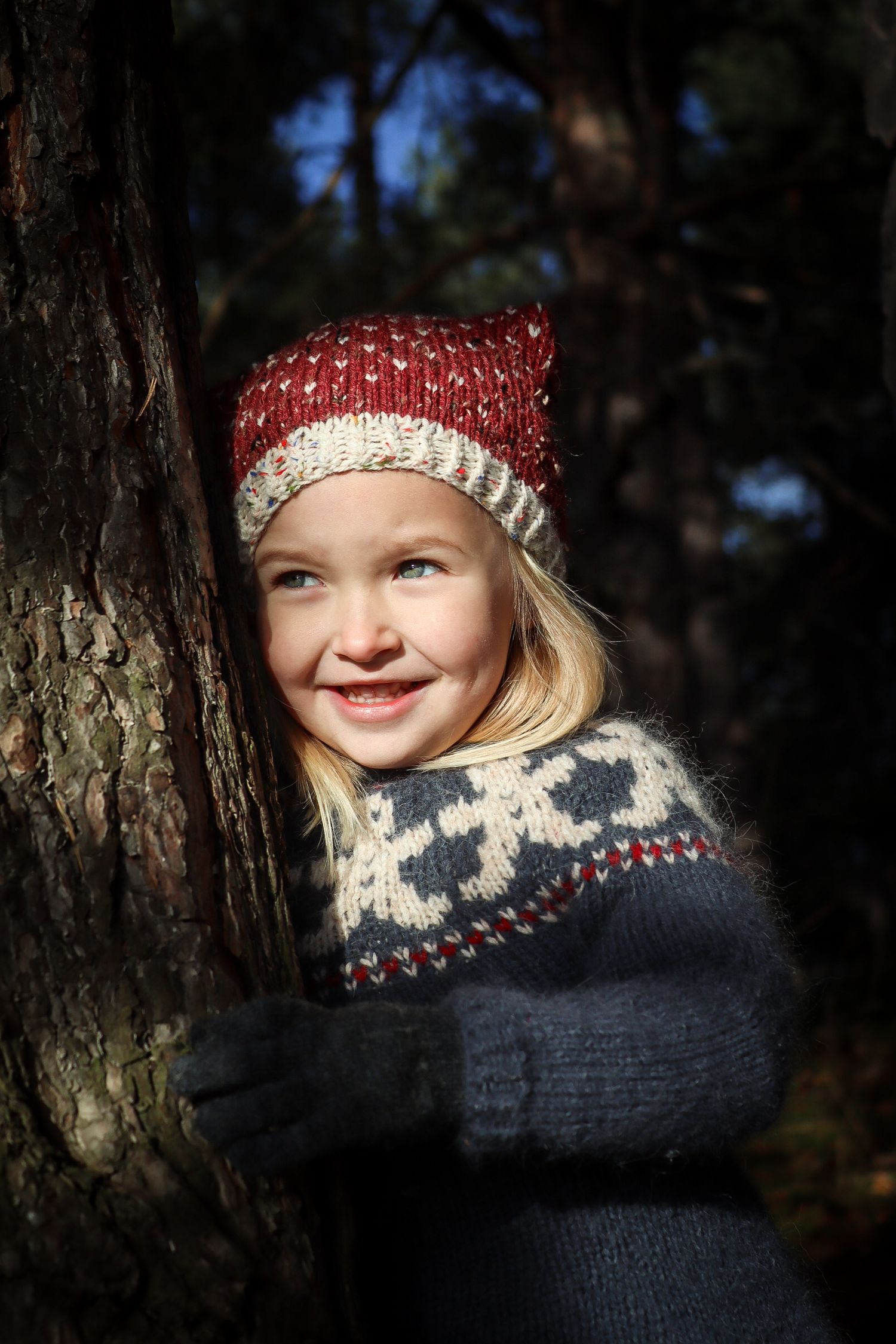 Jente i rød strikkelue og nordisk mønstret genser fotografert ved et tre i skogen.