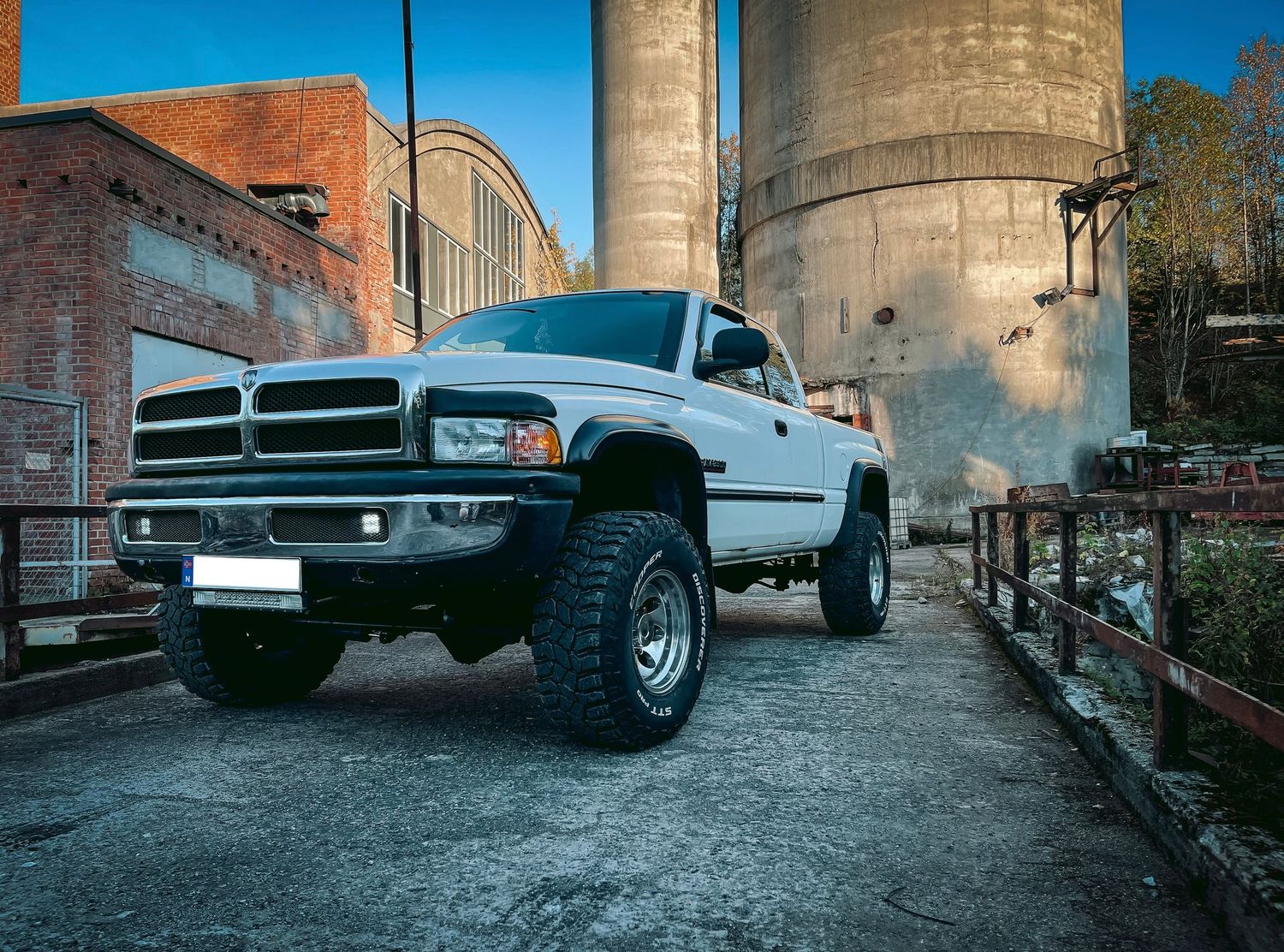 Hvit Dodge Ram pickup fotografert ved industrielle betongsiloer og murbygninger.