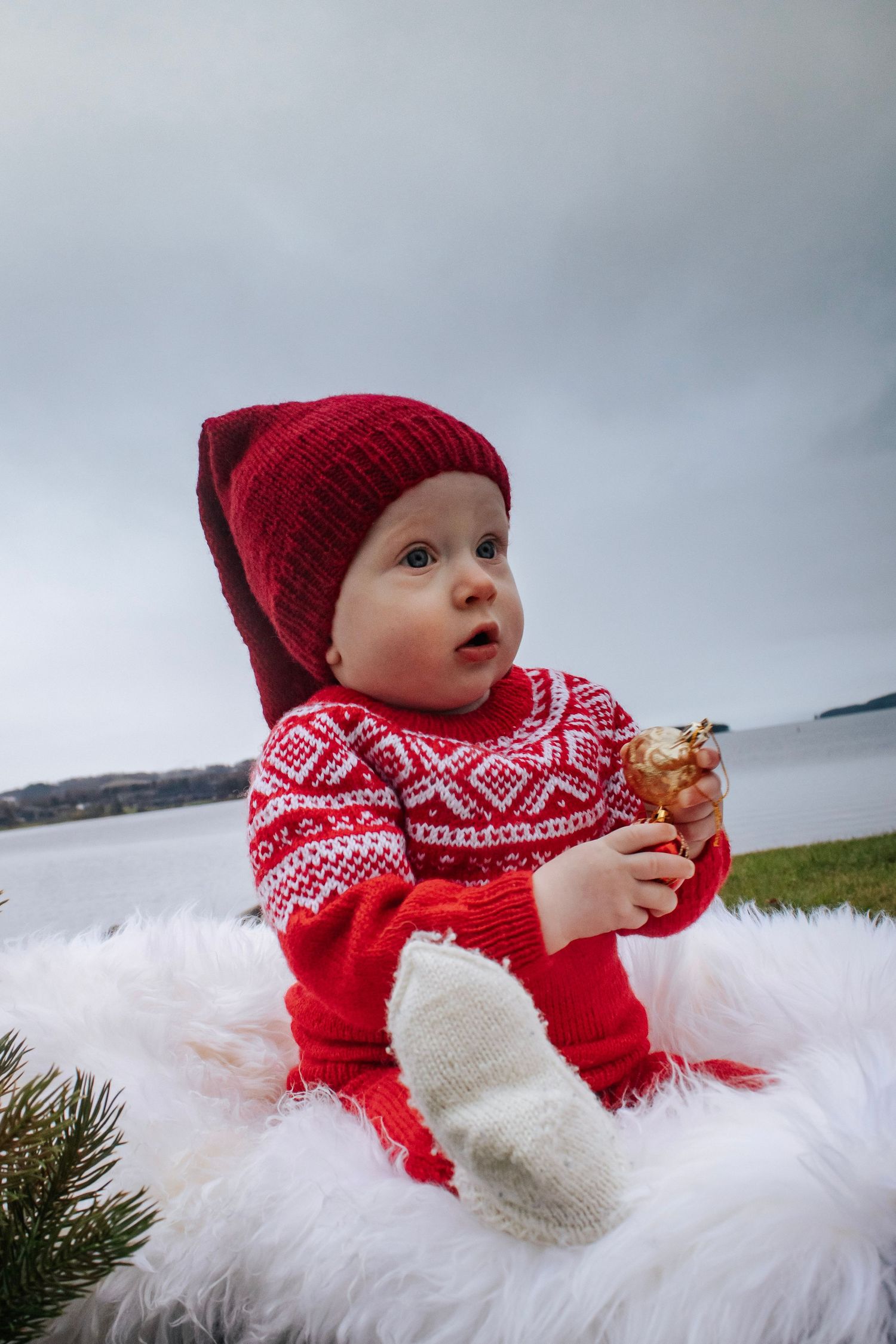 Baby i rød strikkegenser og nisselue fotografert utendørs ved vannet.