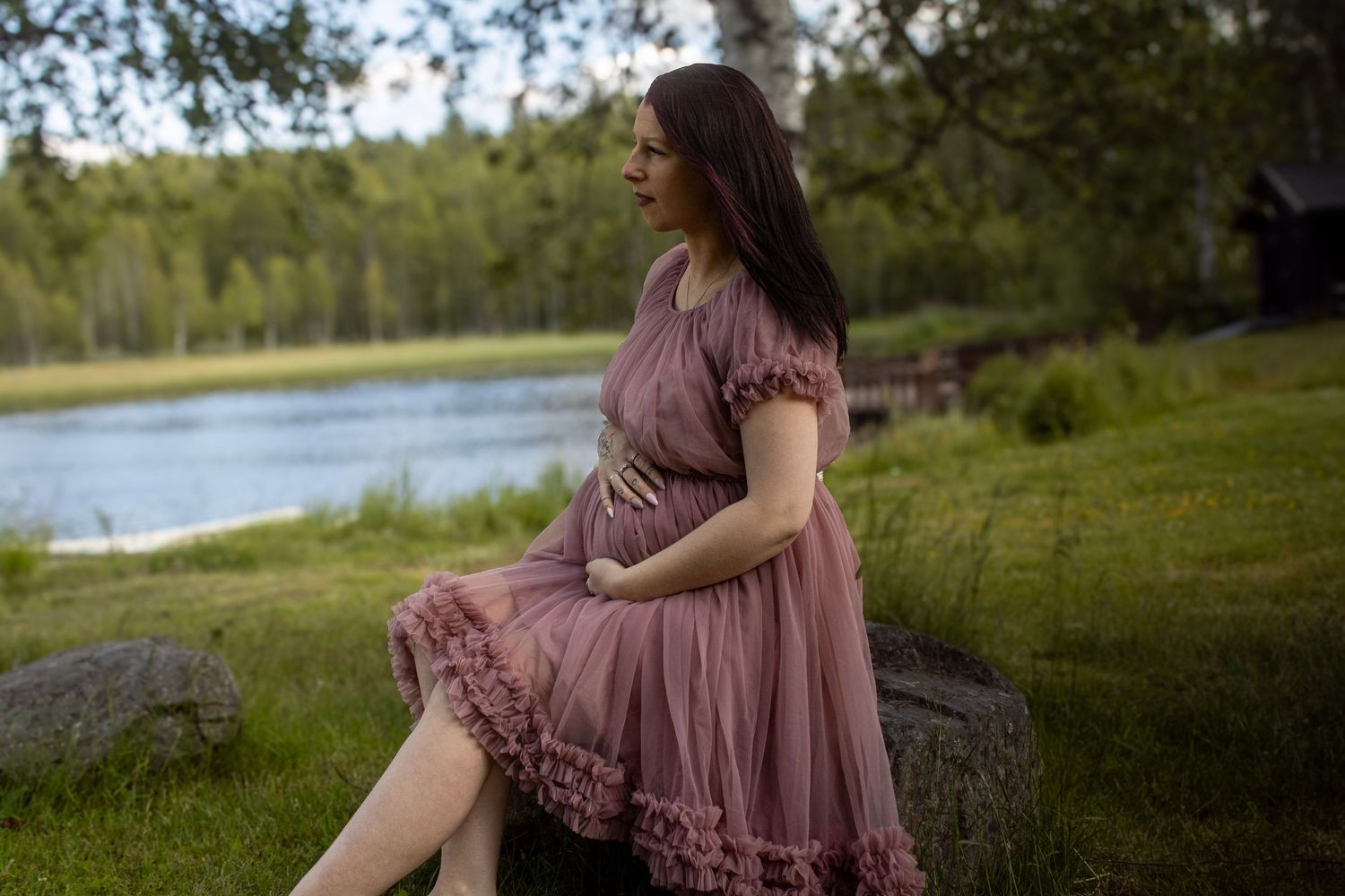 Gravid kvinne i støvrosa kjole sittende på en stein ved Næra i mykt kveldslys.