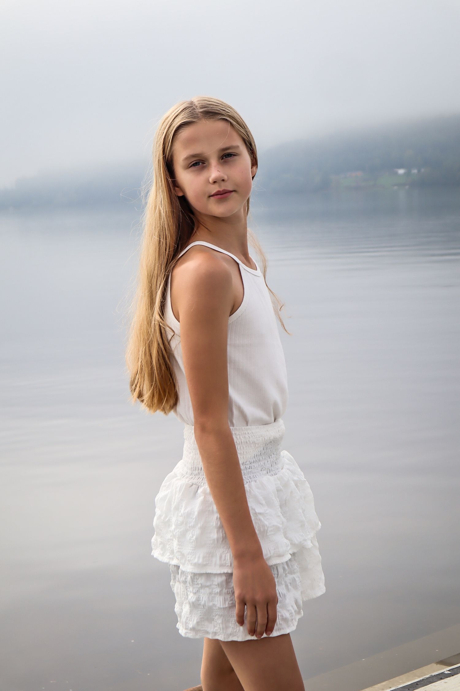 Jente i hvitt antrekk fotografert på en brygge ved et tåkete vann.