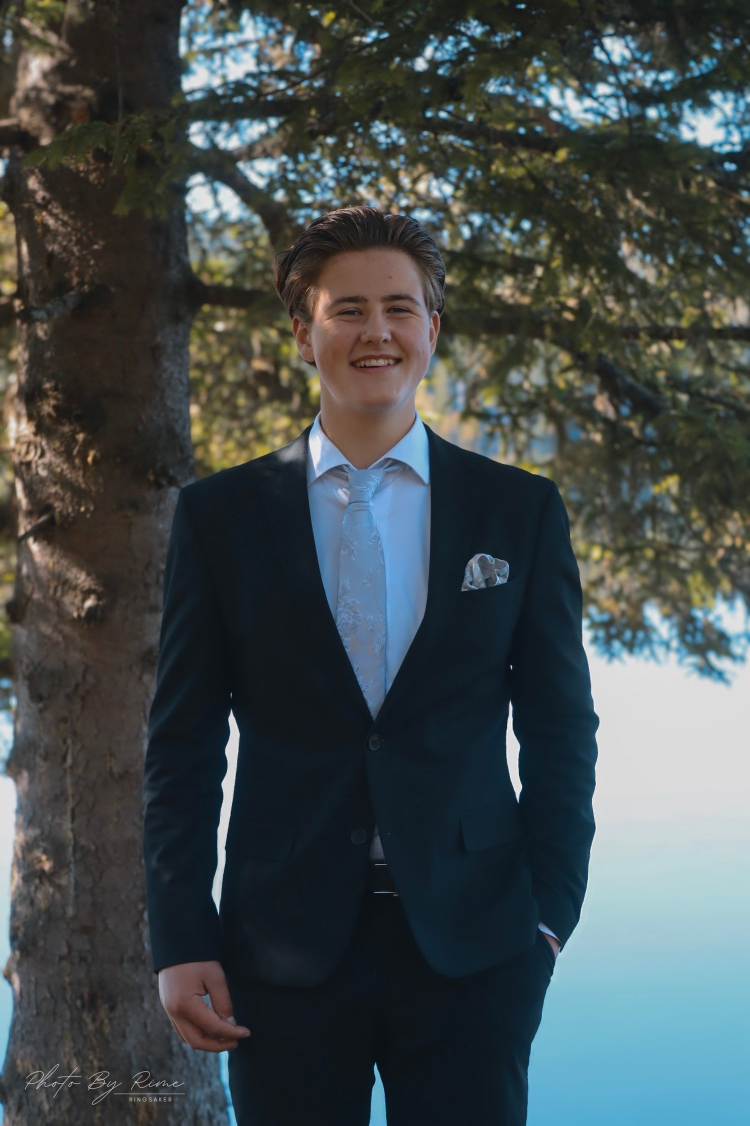 Leende konfirmant i mørk dress fotografert utendørs ved Skumsjøen i Gjøvik.