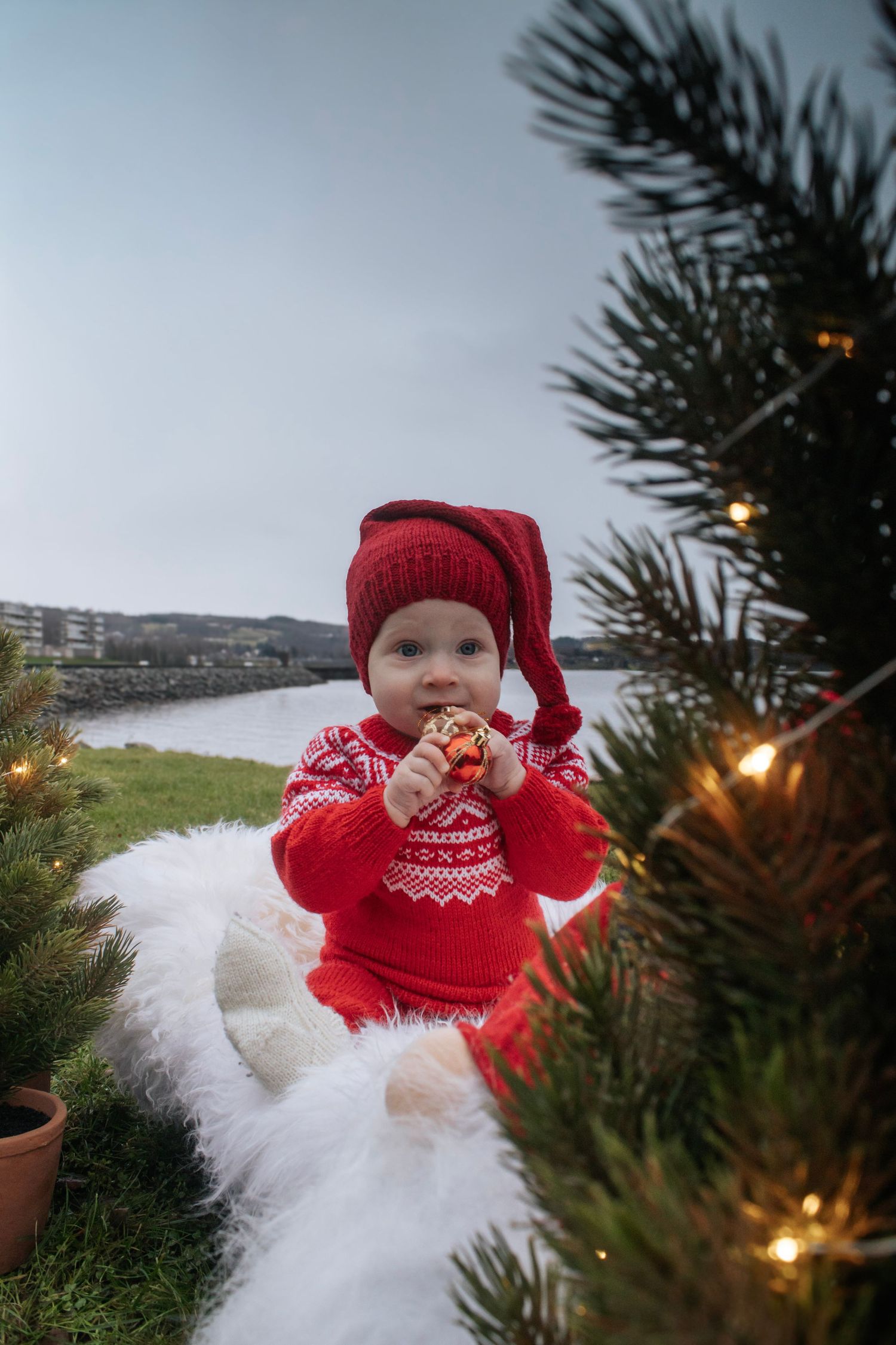 Baby i rød strikkegenser og nisselue sittende på hvitt skinn ved et juletre med lys.