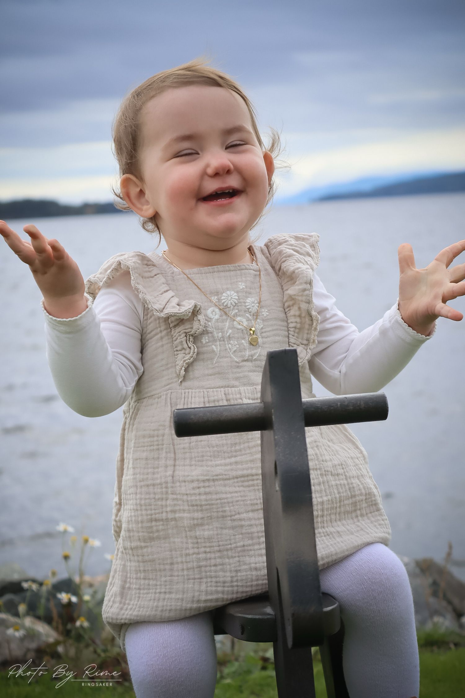 Leende babyjente i beige kjole fotografert ved vannet.