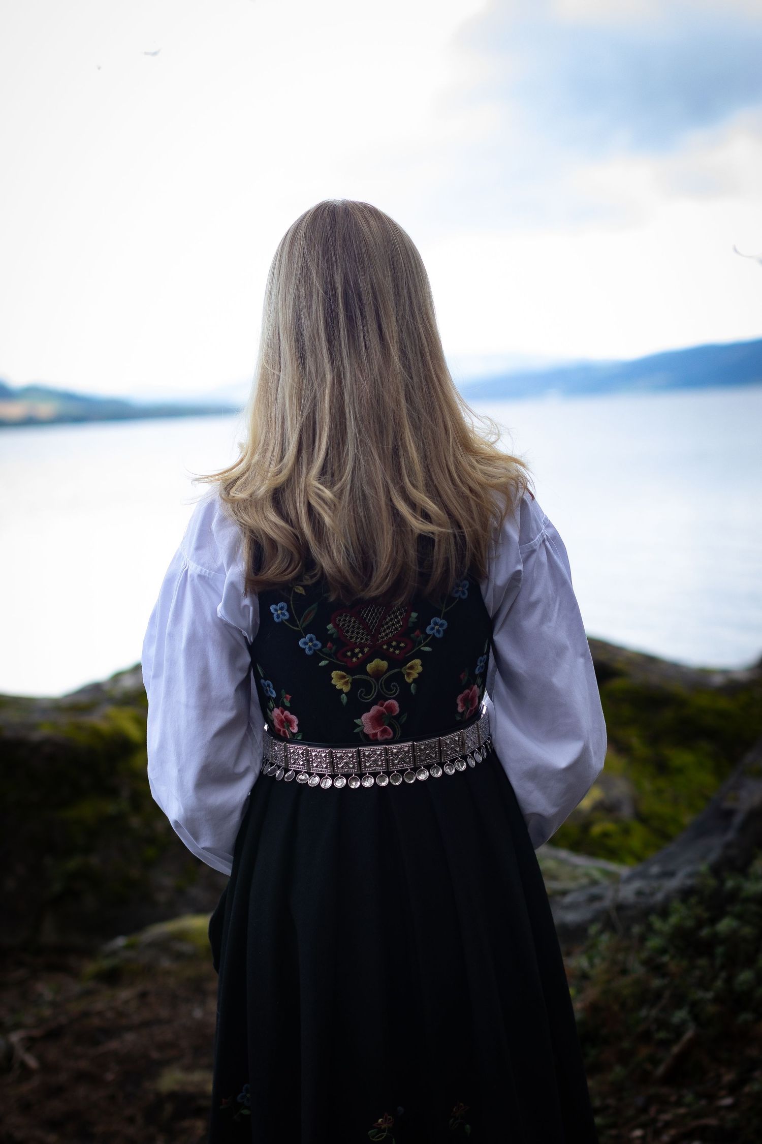 Konfirmant i brodert bunad fotografert bakfra ved vannet.