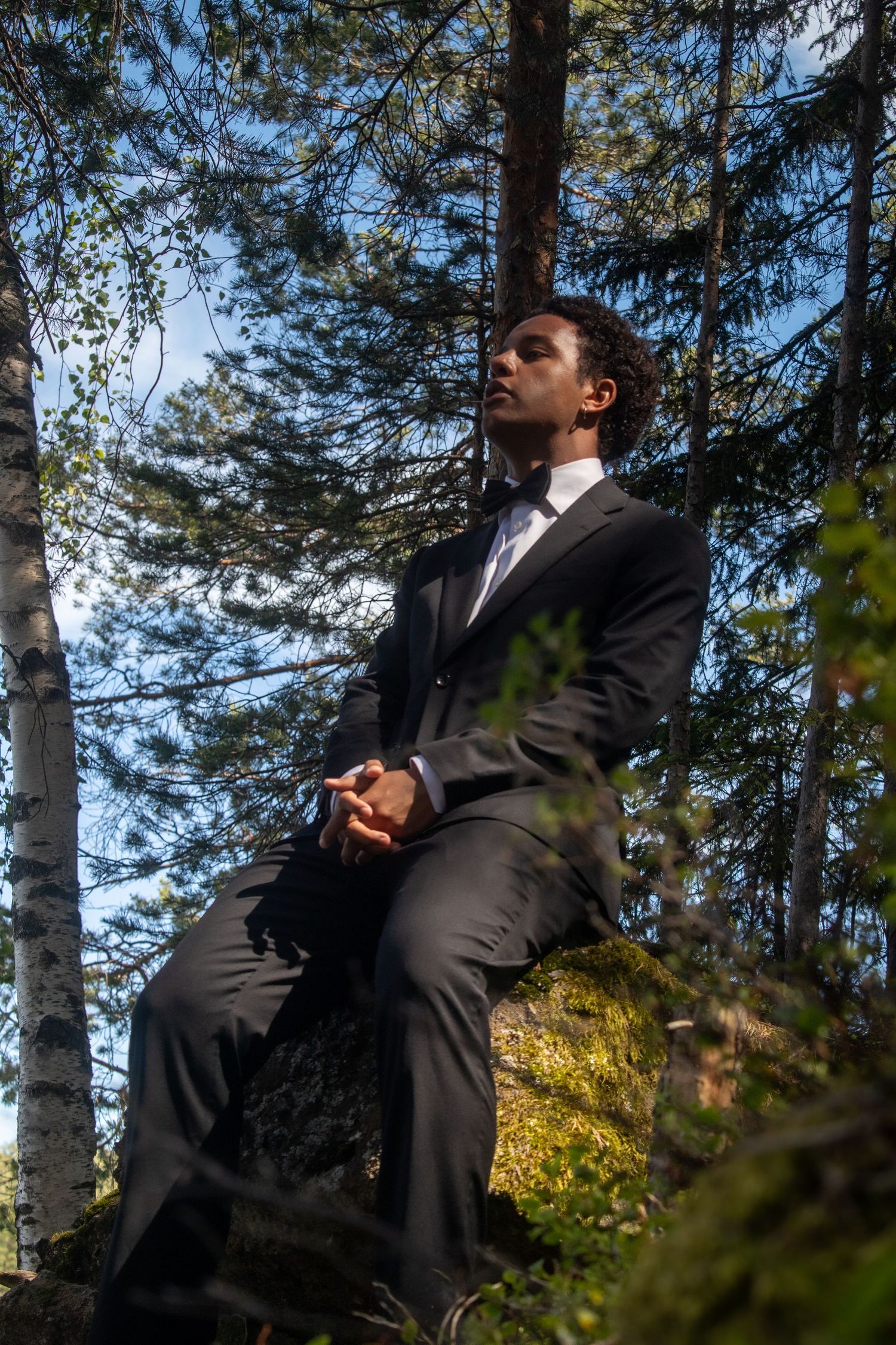 Mann i svart dress sittende på en mosegrodd stein i en solfylt skog.