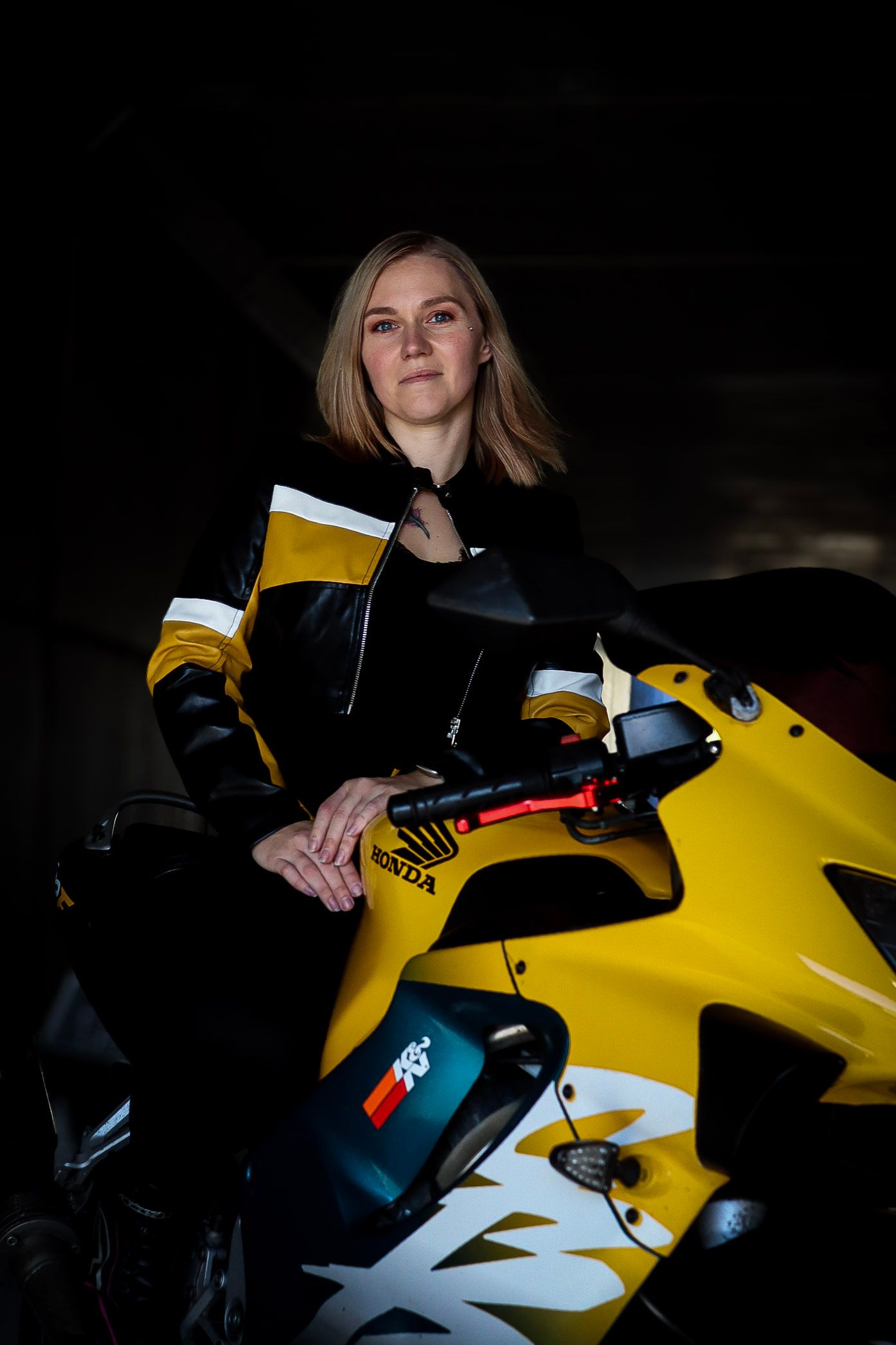 Kvinne i svart og gul skinnjakke fotografert med en gul Honda motorsykkel.