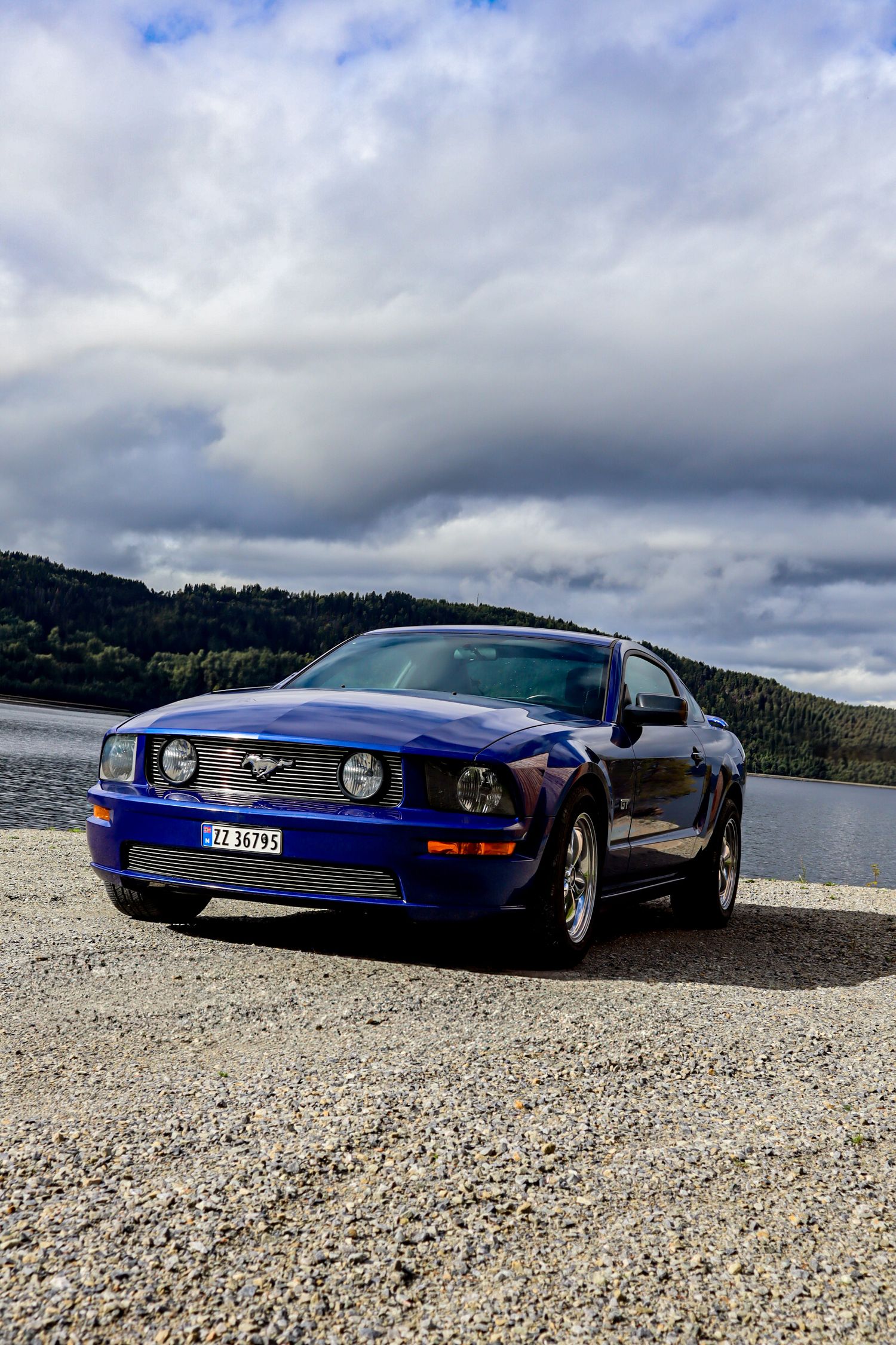 Blå Ford Mustang GT fotografert på steinete underlag ved Mjøsa under overskyet himmel.