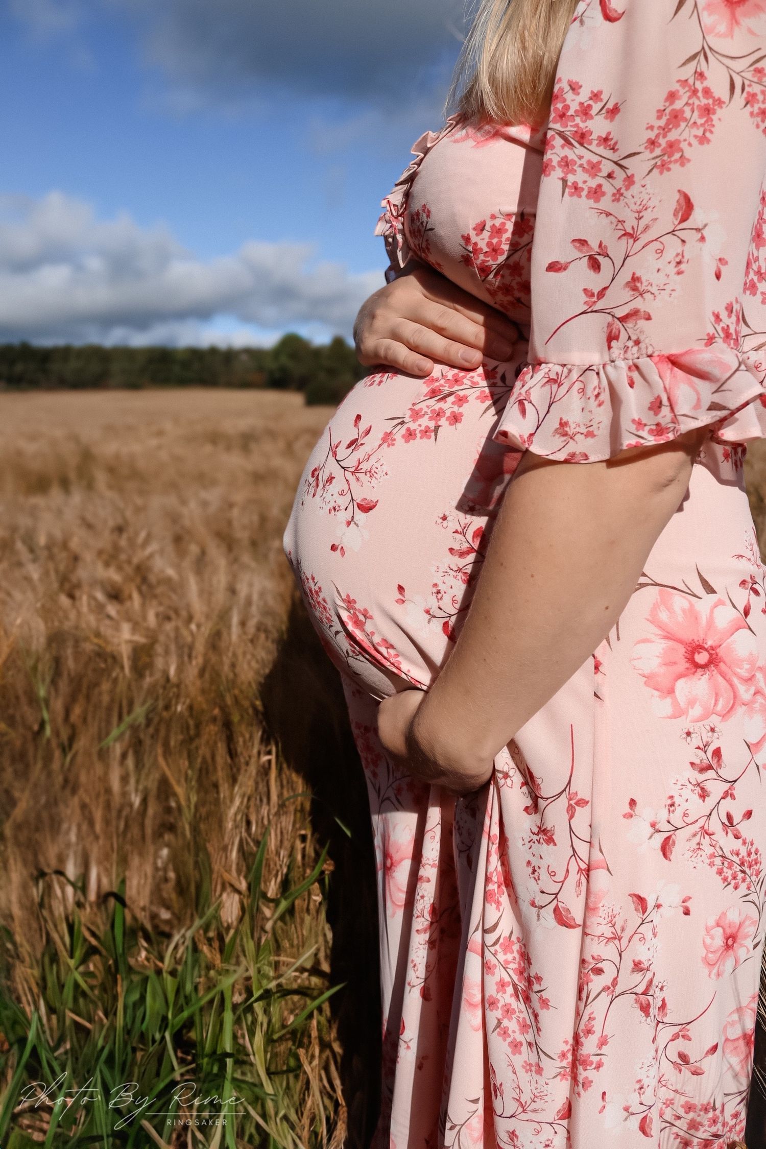 Nærbilde av gravid kvinne i rosa blomsterkjole med hendene rundt gravidmagen i en kornåker.