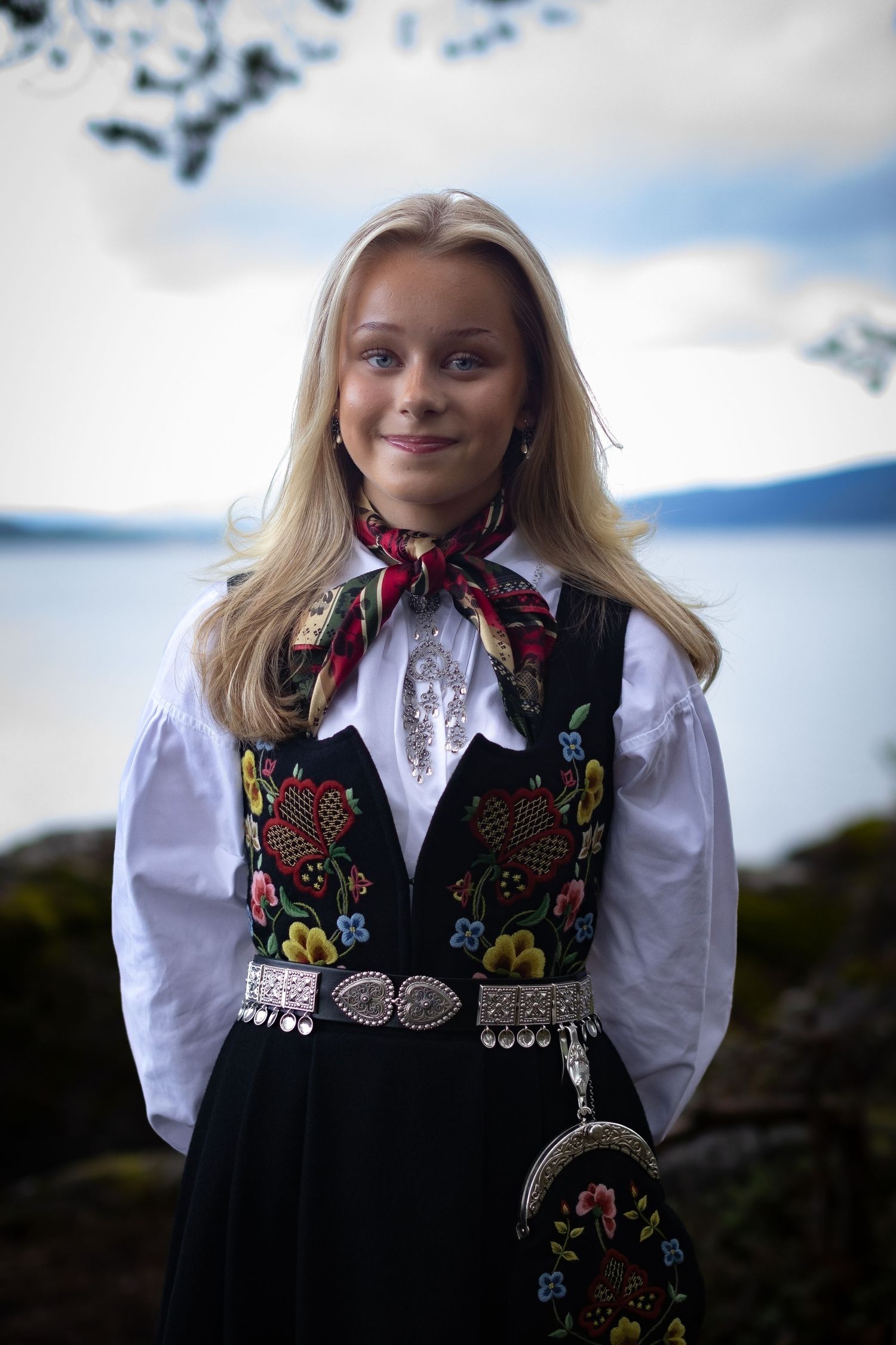 Konfirmant i tradisjonell norsk bunad med sølvbelte fotografert ved vannet.