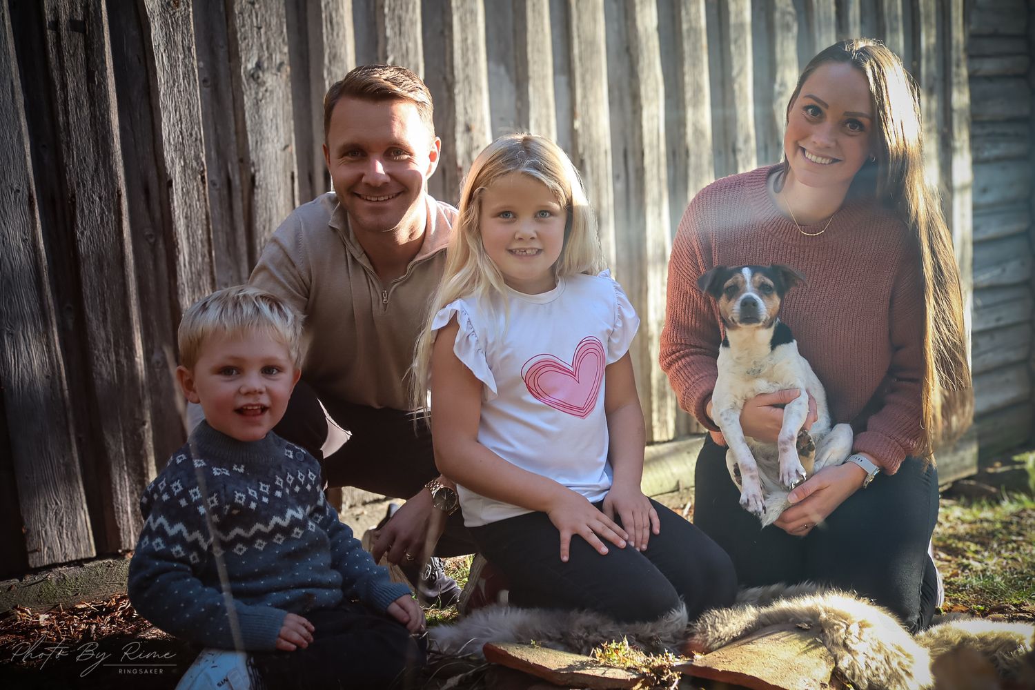 Familie med to barn og hund fotografert sammen ved et rustikt tregjerde.