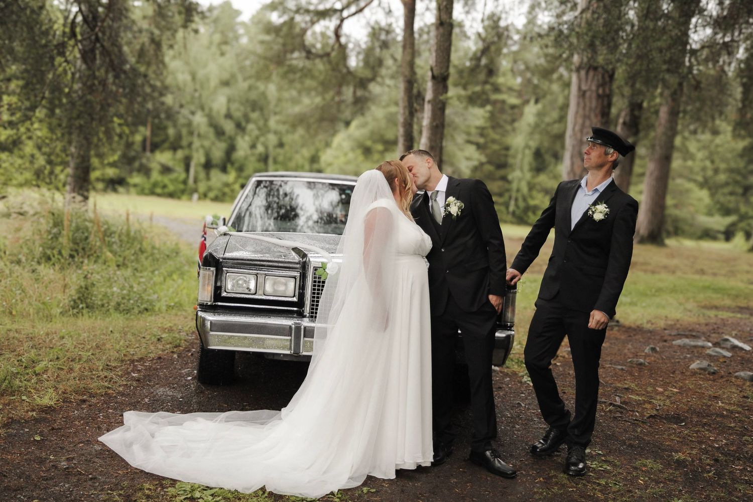 Brudepar som kysser ved en hvit veteranbil under bryllupsfotografering i skogen.