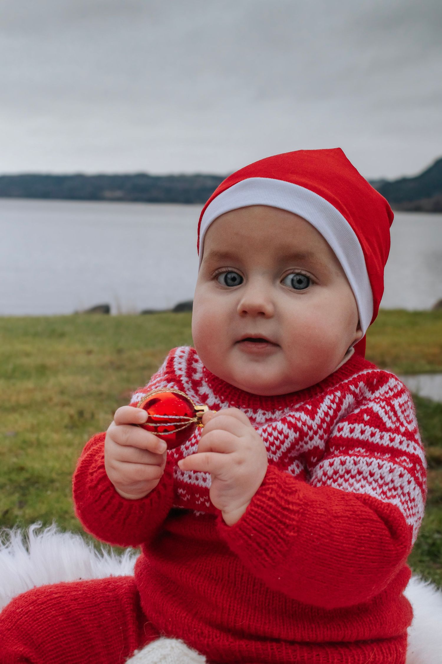 Baby i rød strikkegenser og nisselue med en rød julekule i hånden, fotografert ved vannet.