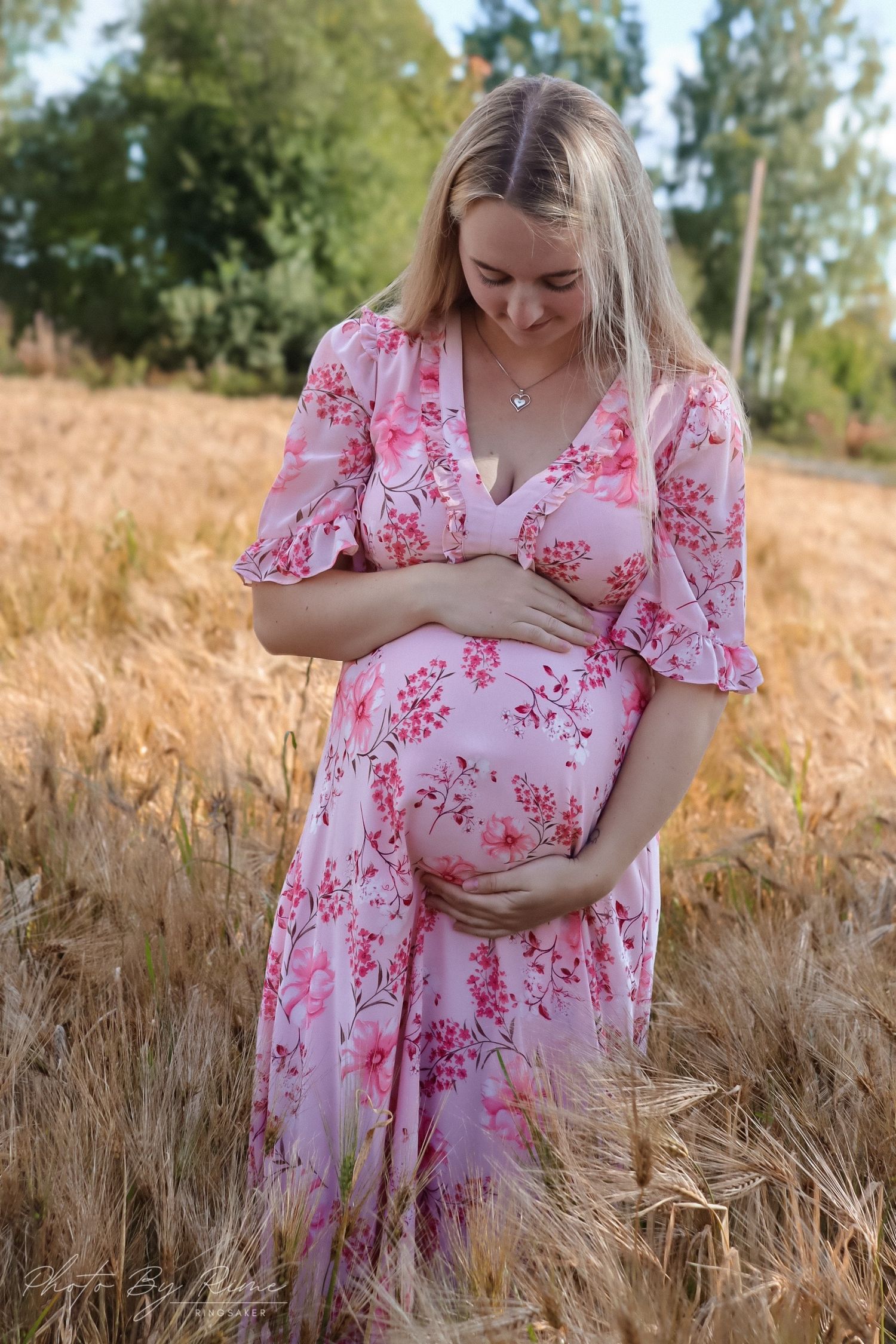 Gravid kvinne i rosa blomsterkjole fotografert i en kornåker.
