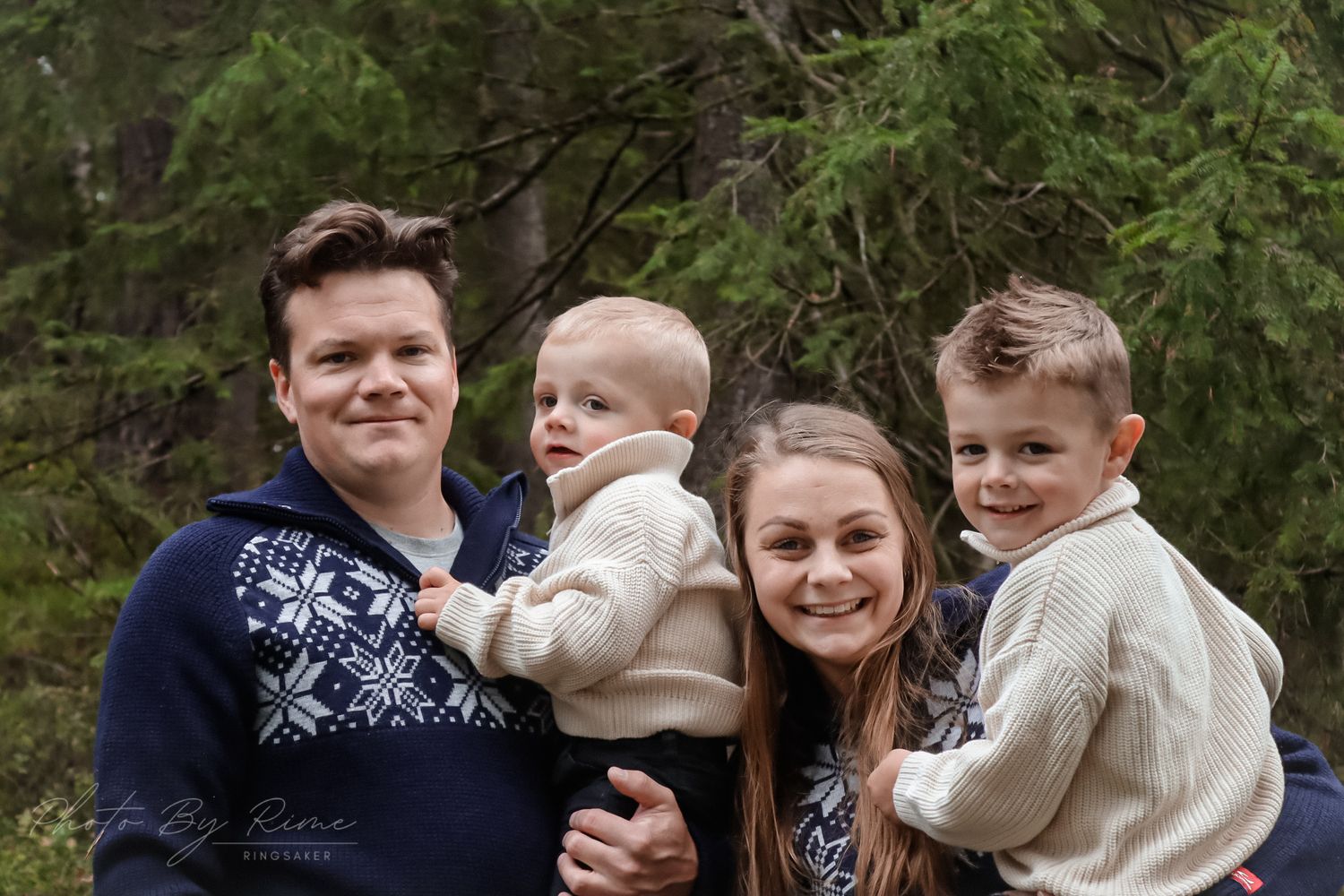 Familie på fire med baby fotografert sammen blant grønne furutrær i skogen.