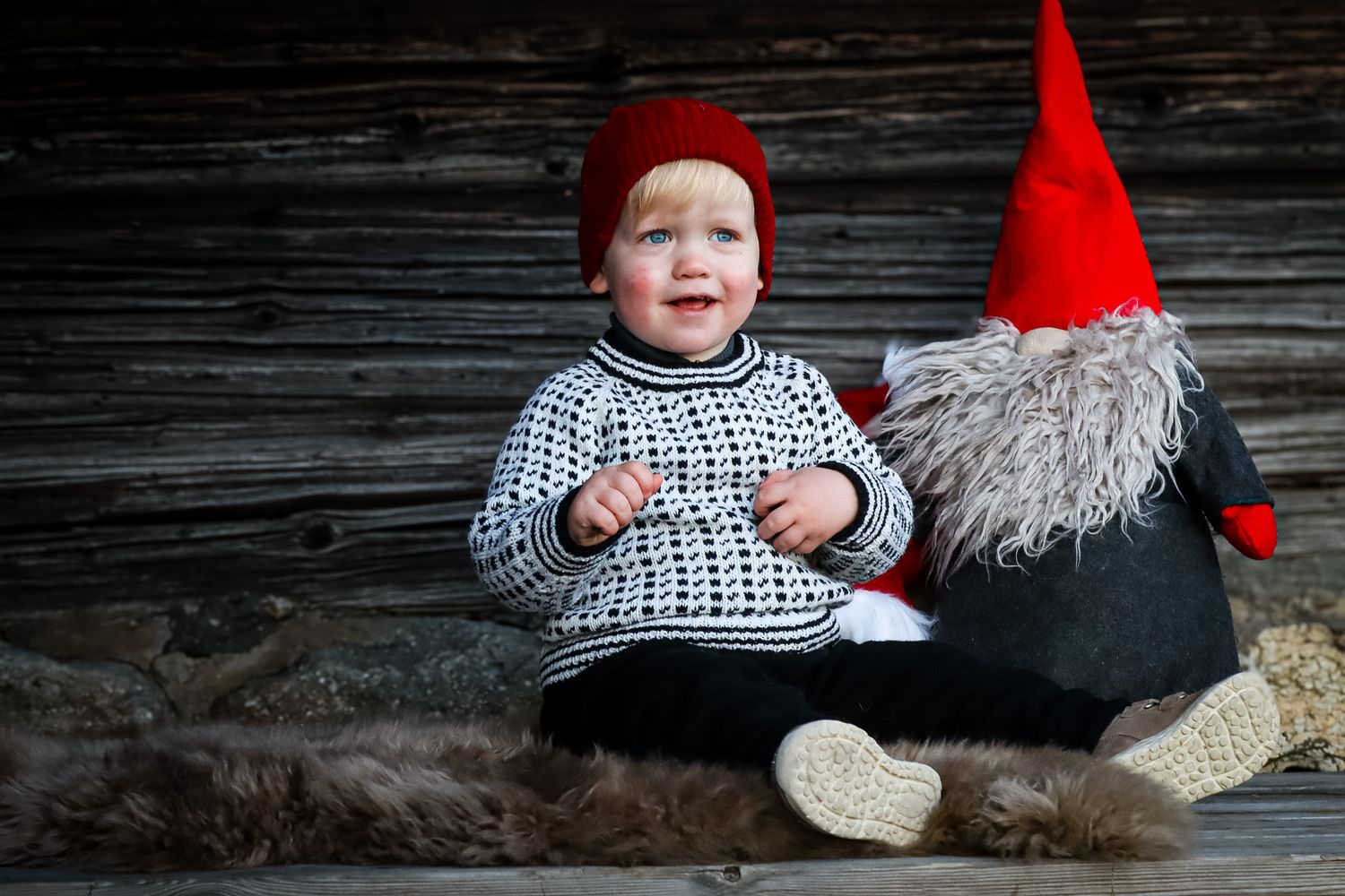 Baby i rød lue og mønstret genser fotografert ved juledekorasjon utendørs.