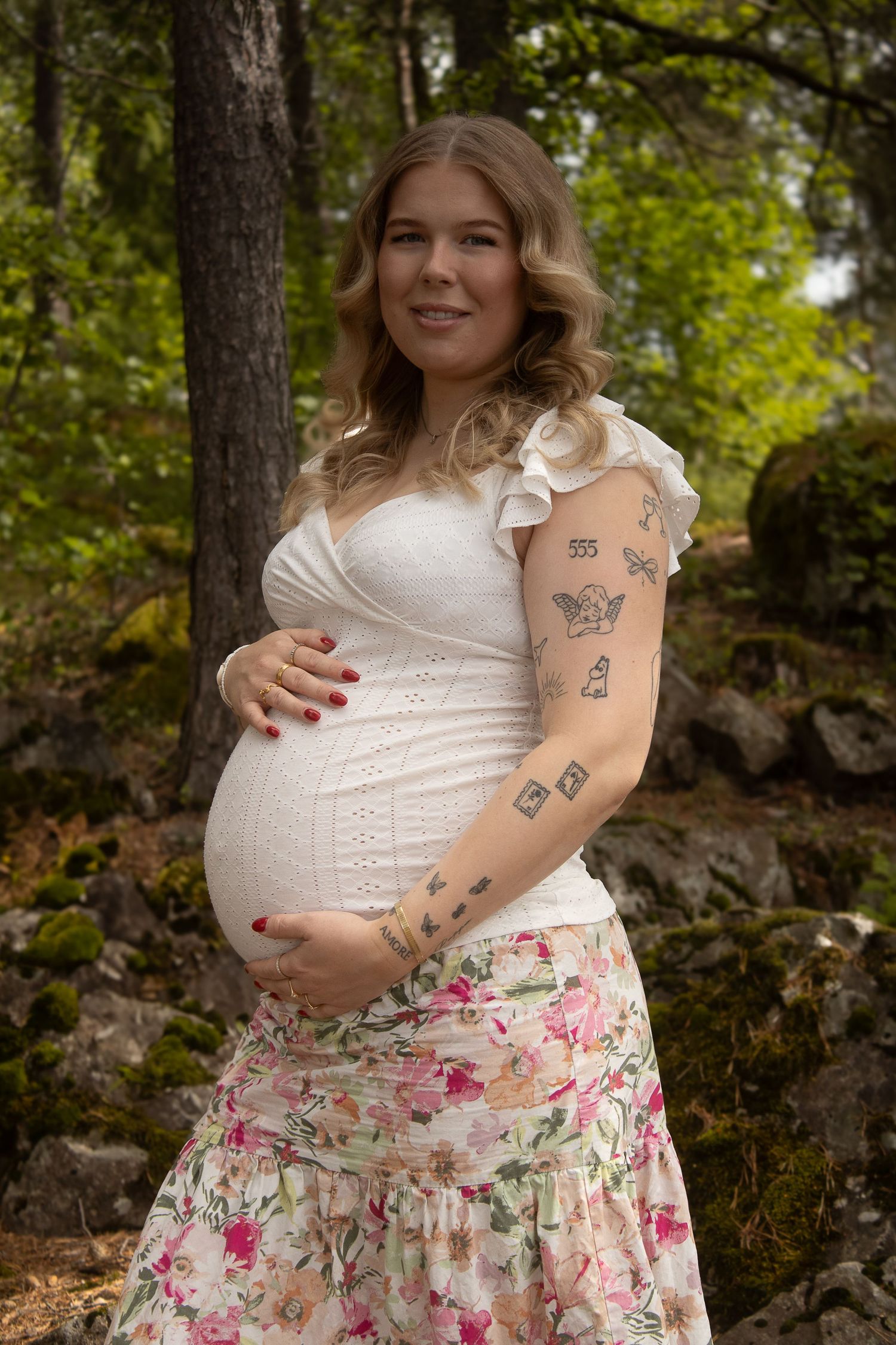 Gravid kvinne i hvit topp og blomstrete skjørt fotografert i grønn skog.