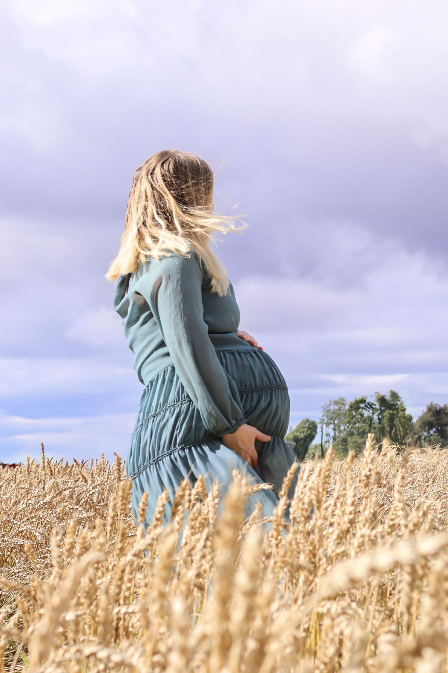 Gravid kvinne i petrolfarget kjole stående i en gyllen kornåker under overskyet himmel.