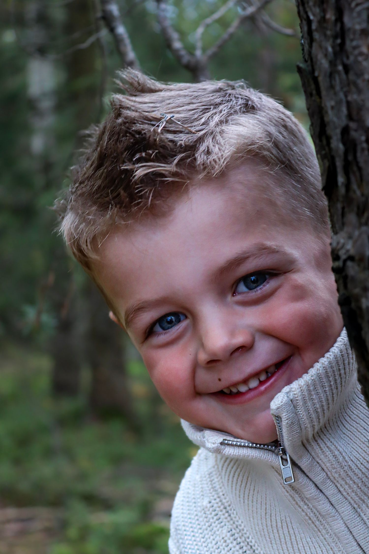 Smilende gutt i lys genser lent mot et tre i skogen.