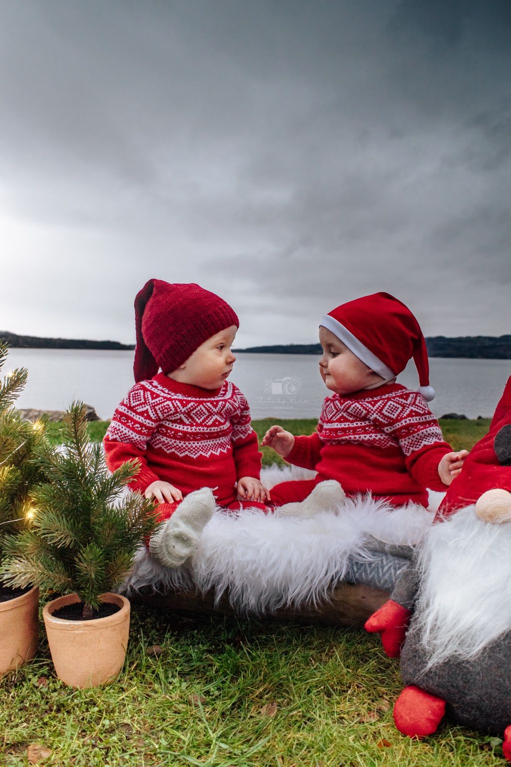 To babyer i matchende røde julegensere og luer fotografert utendørs ved vannet.