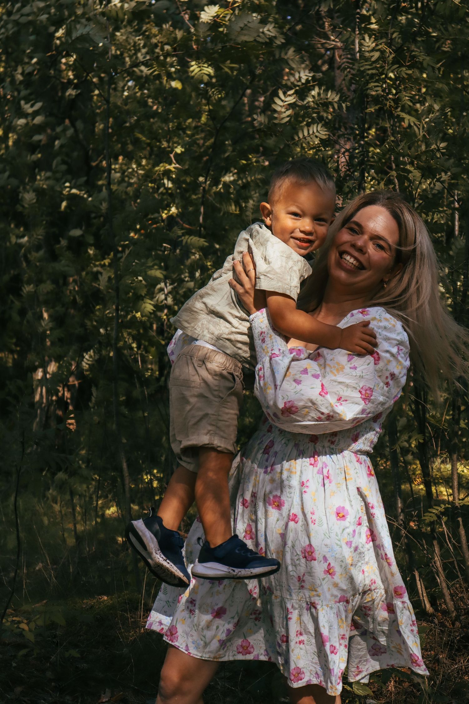 Mor i blomstrete kjole med en leende liten gutt på armen under familiefotografering i skogen.