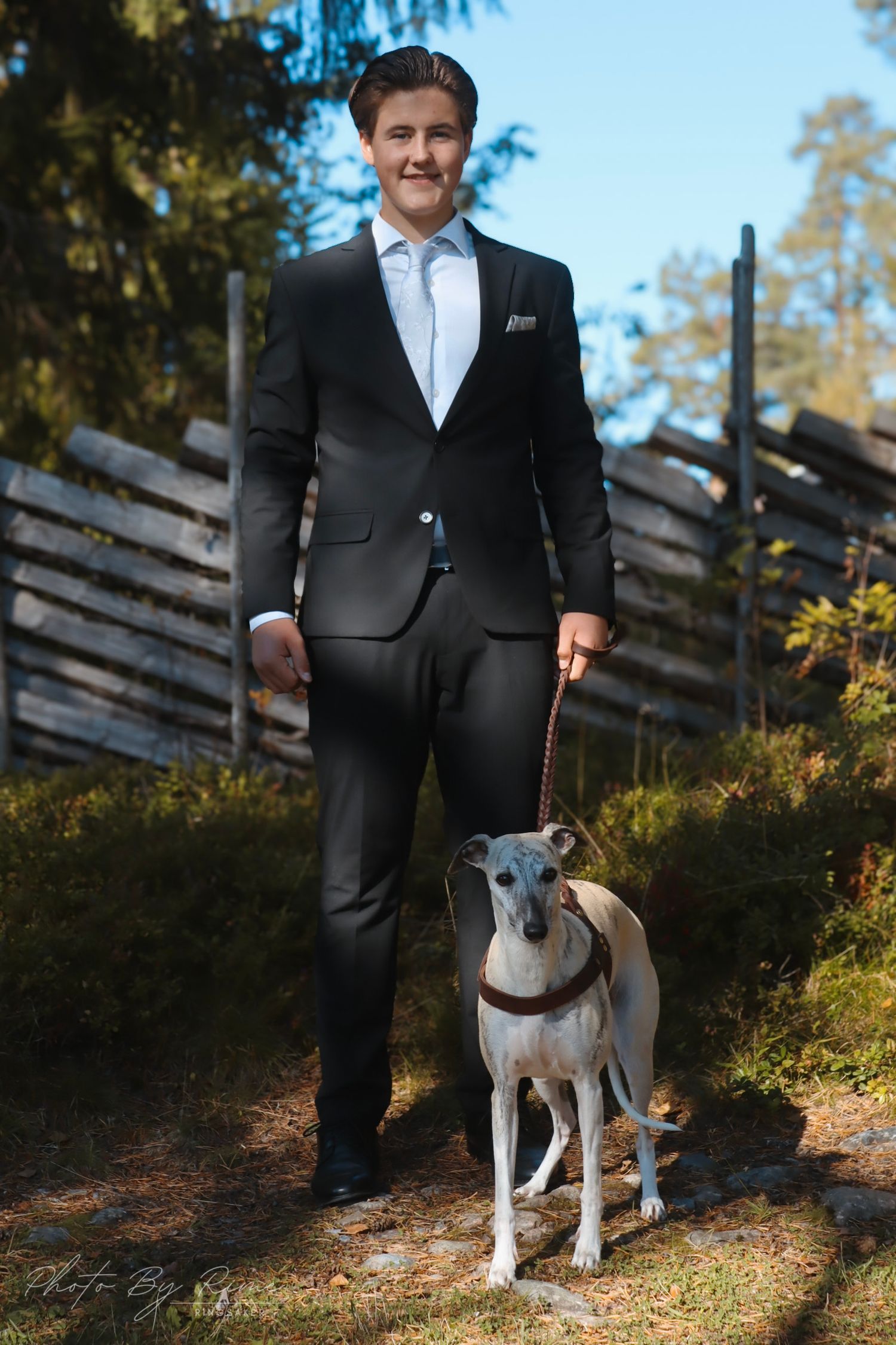 Konfirmant i svart dress fotografert sammen med en hvit Whippet utendørs.