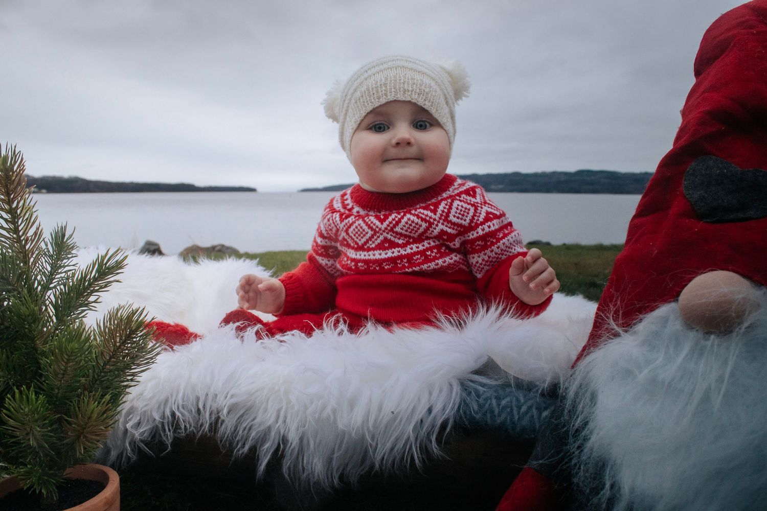 Baby i rød strikkegenser og hvit lue sittende på hvitt skinn utendørs ved vannet.