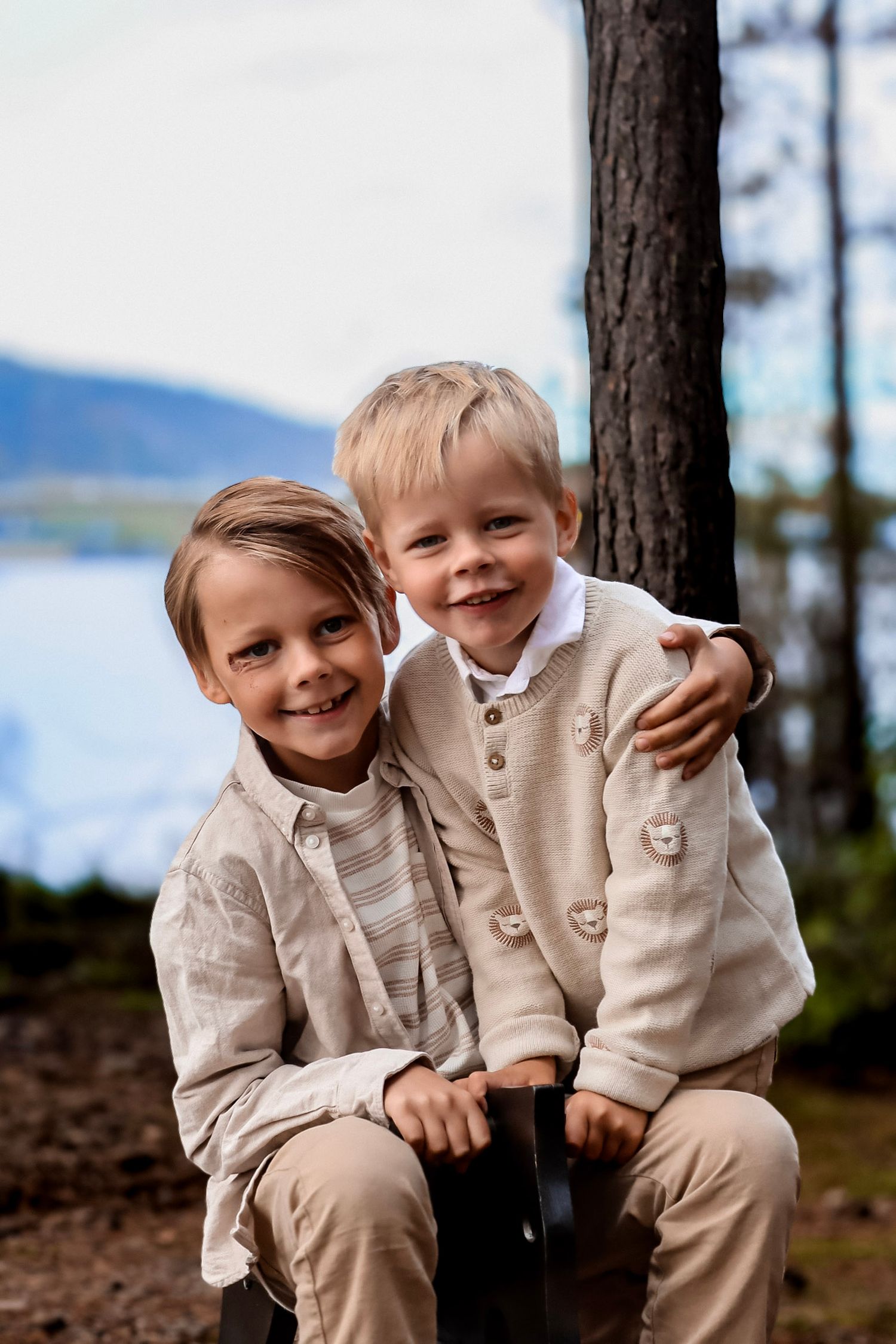 To brødre som smiler og klemmer hverandre ved et tre med vann i bakgrunnen.