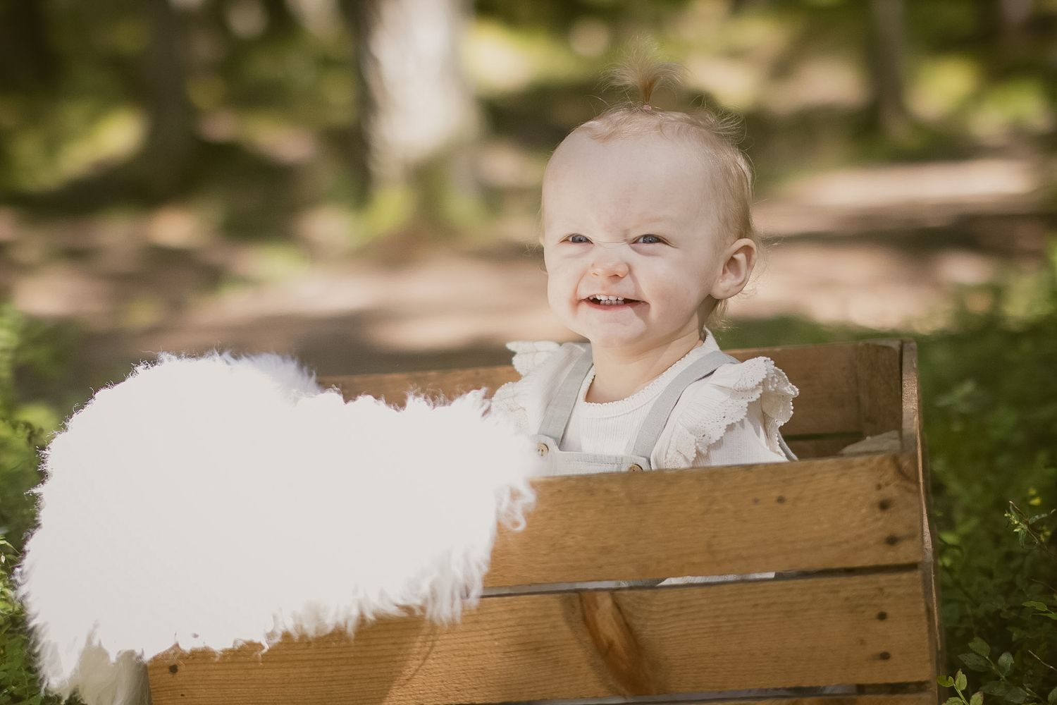Smilende baby i hvite klær sittende i en trekasse utendørs.