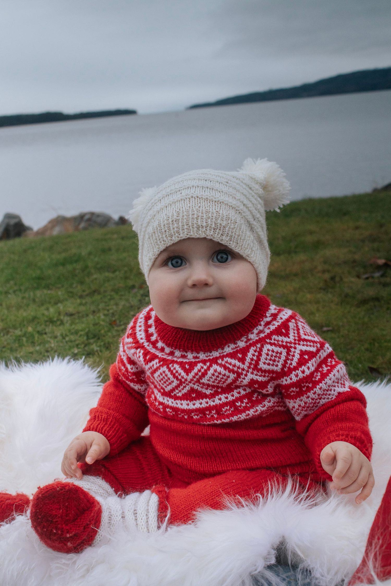 Baby i rød strikkegenser og hvit lue sittende på hvitt skinn utendørs ved vannet.