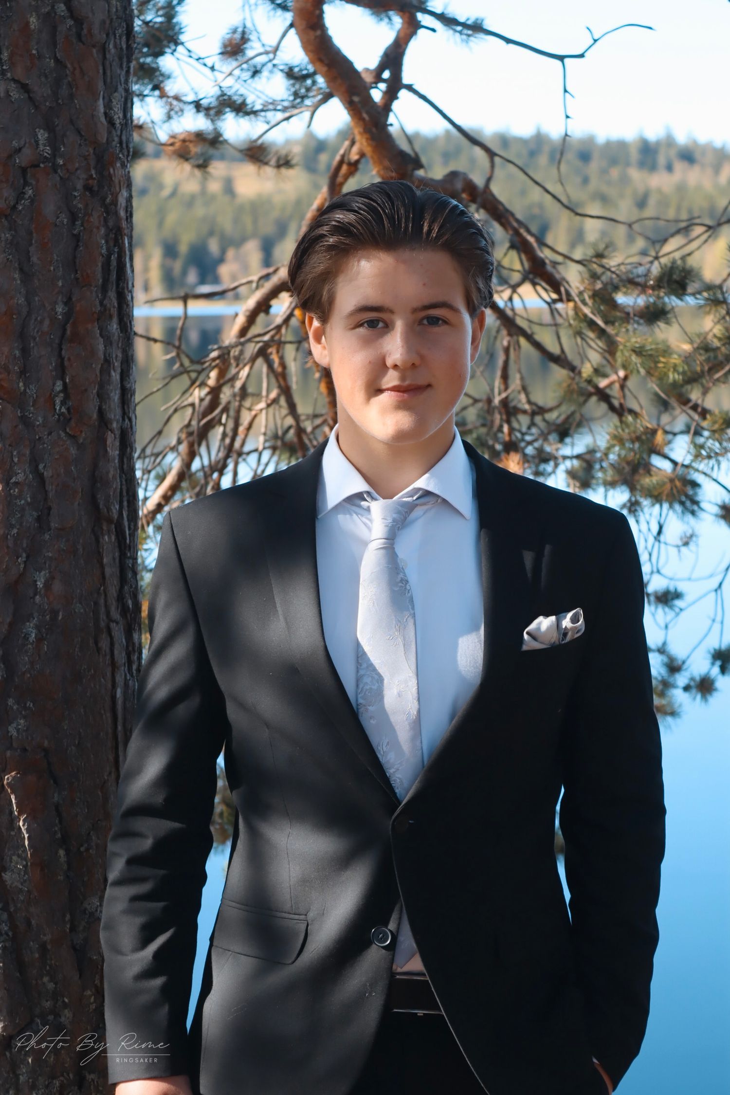 Konfirmant i svart dress lent mot et furutre ved Skumsjøen i Gjøvik.