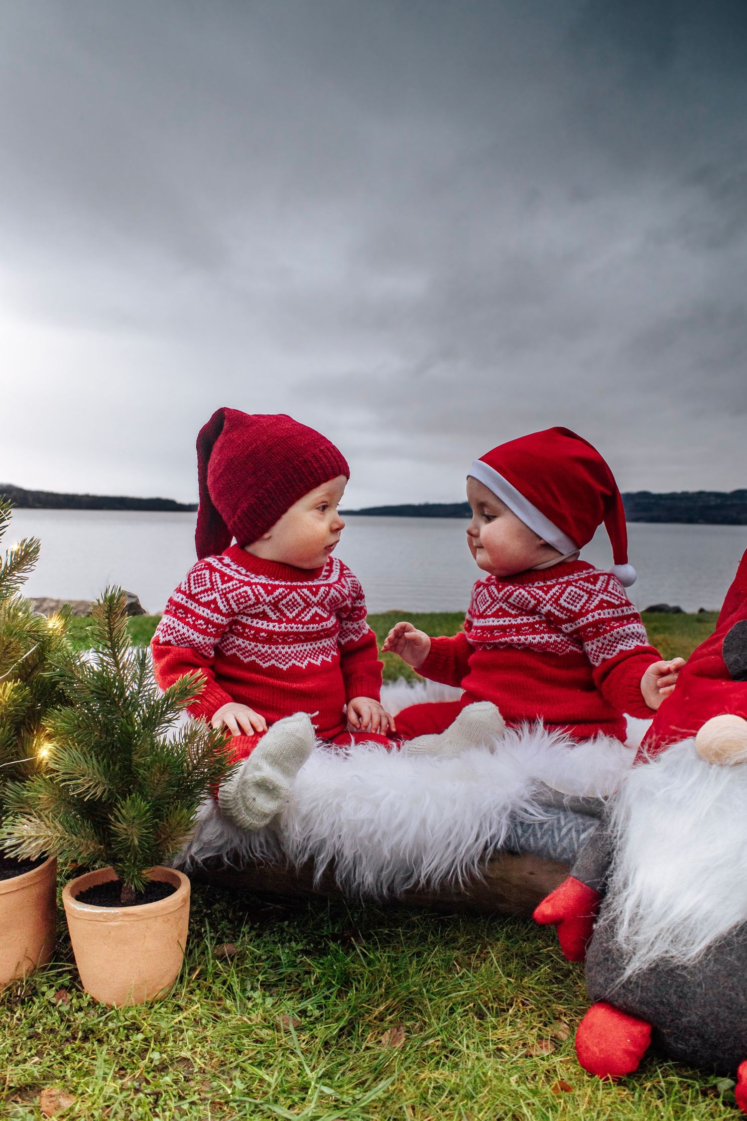To babyer i matchende røde strikkegensere og nisseluer fotografert sammen utendørs ved vannet.