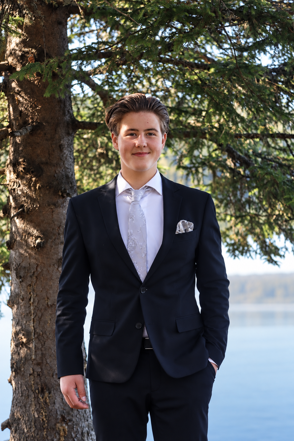 Konfirmant i mørk dress fotografert utendørs ved Skumsjøen i Gjøvik, med furutrær og vann i bakgrunnen.