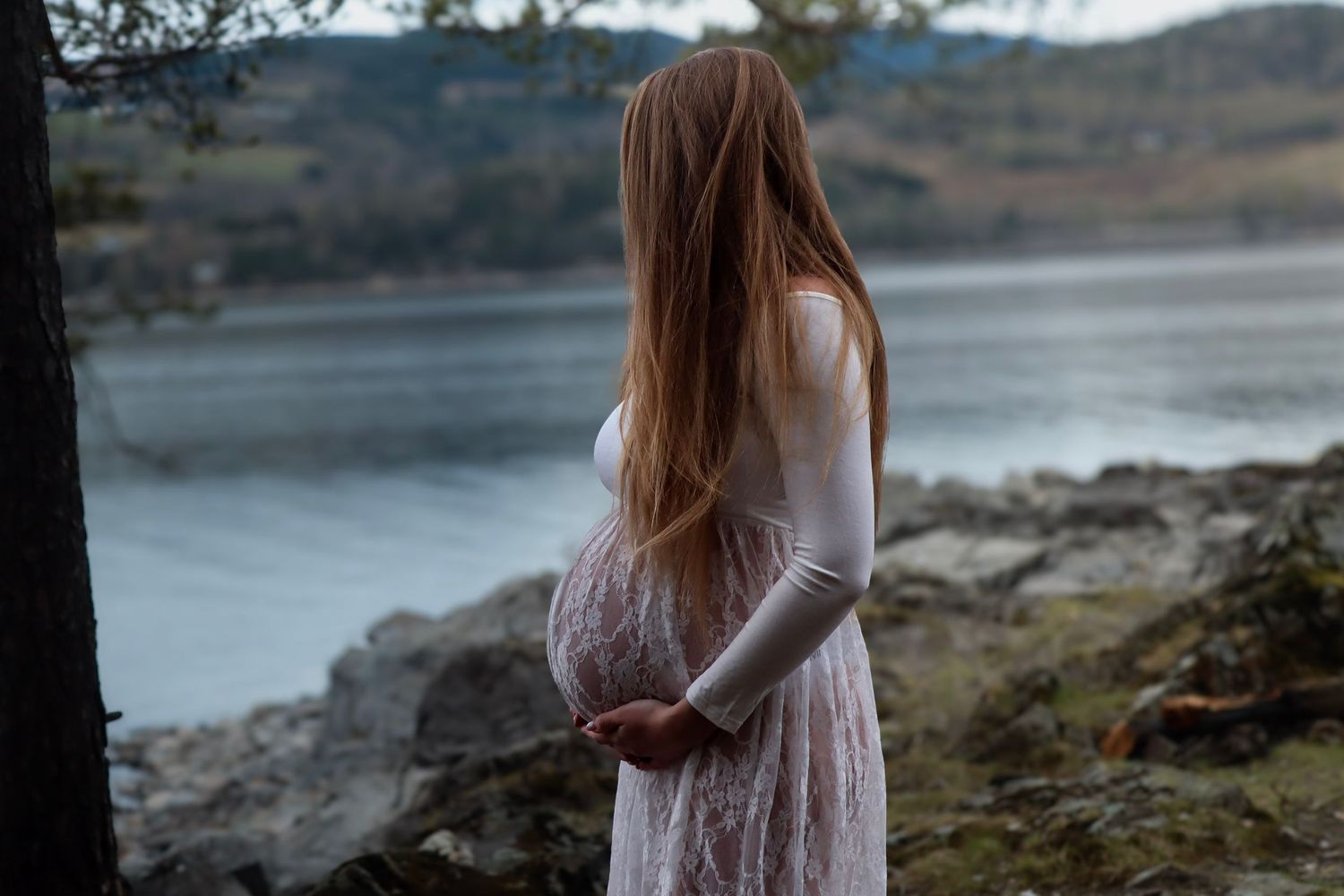 Gravid kvinne i hvit blondekjole fotografert ved vannet.